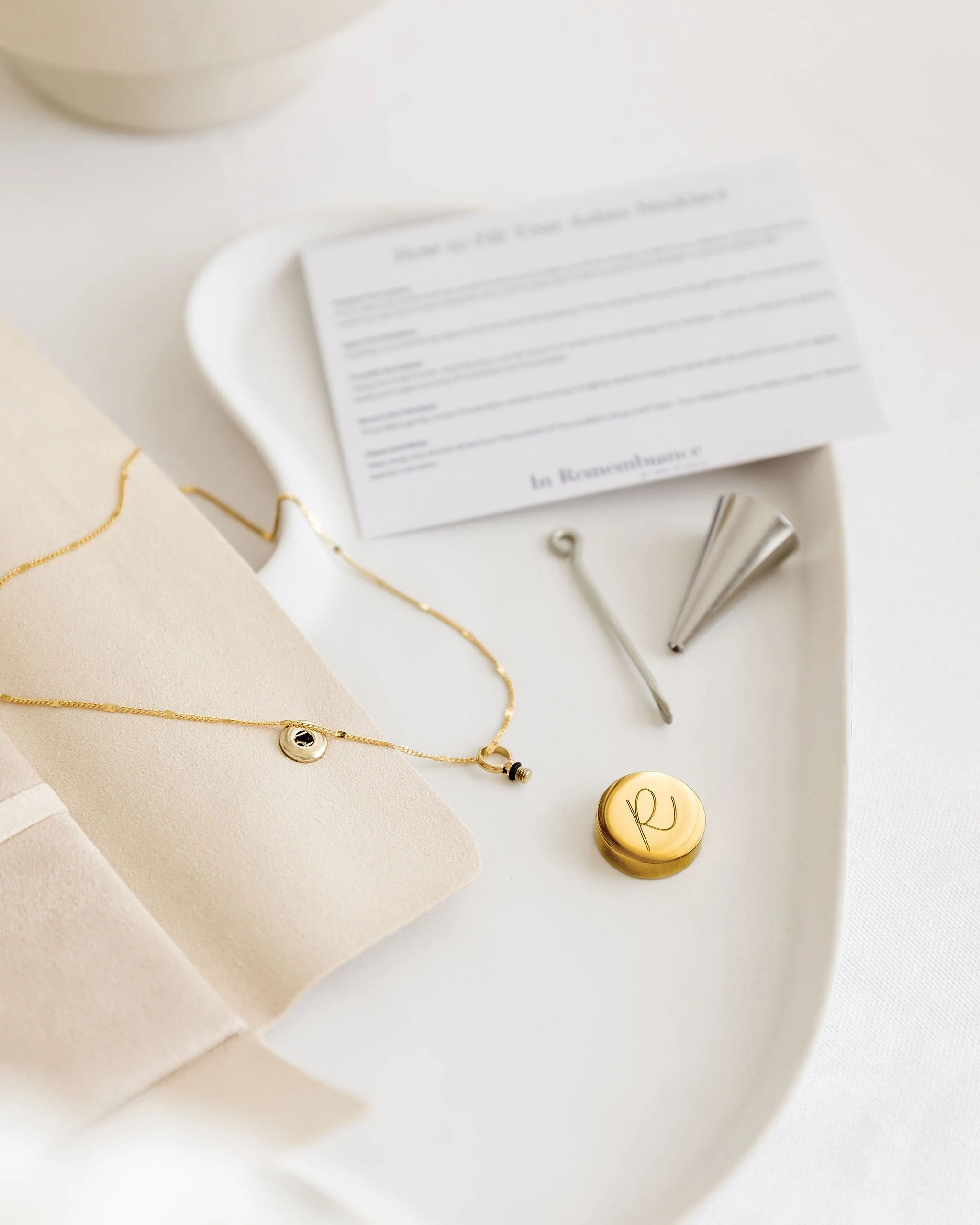 Jewelry and accessories on a white tray, including a gold necklace with a round pendant, a twist wine opener, a coffee tamper, a gold container with an engraved letter, and a partially visible white cup on a beige fabric background.