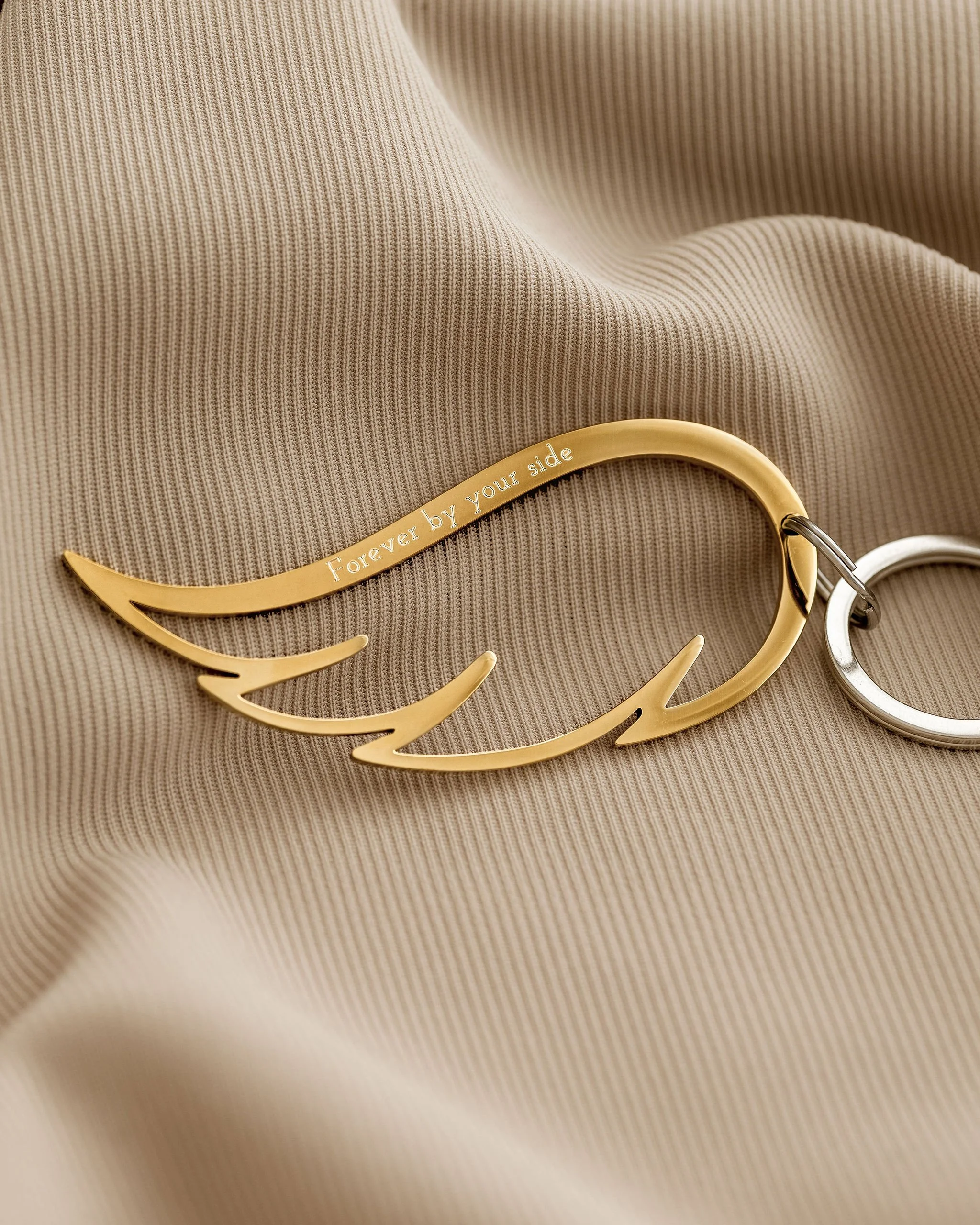 Gold keychain shaped like angel wings with the inscription "Forever by your side," placed on beige fabric.