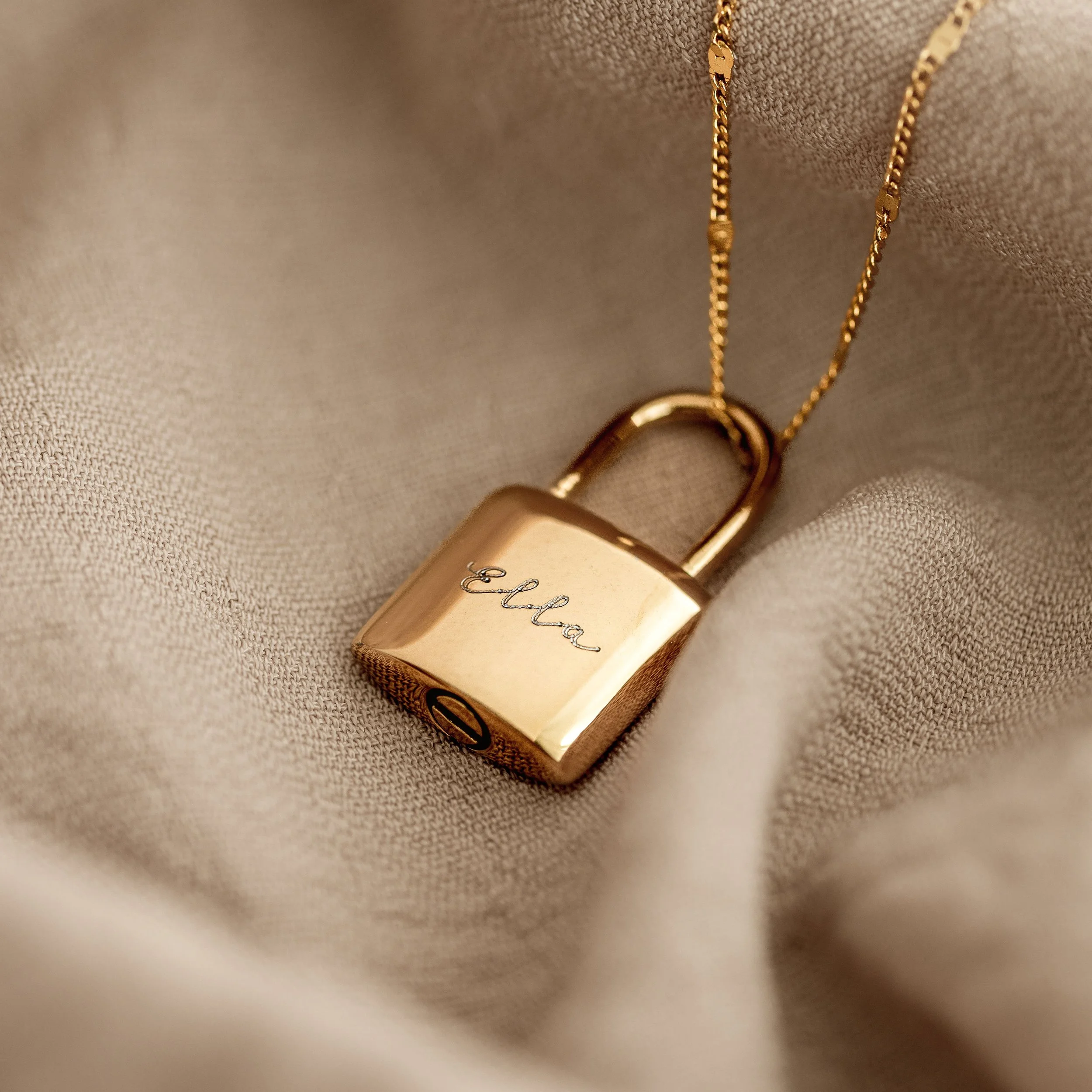 Gold lock pendant necklace with the name 'Eleanor' engraved on it, resting on beige fabric.