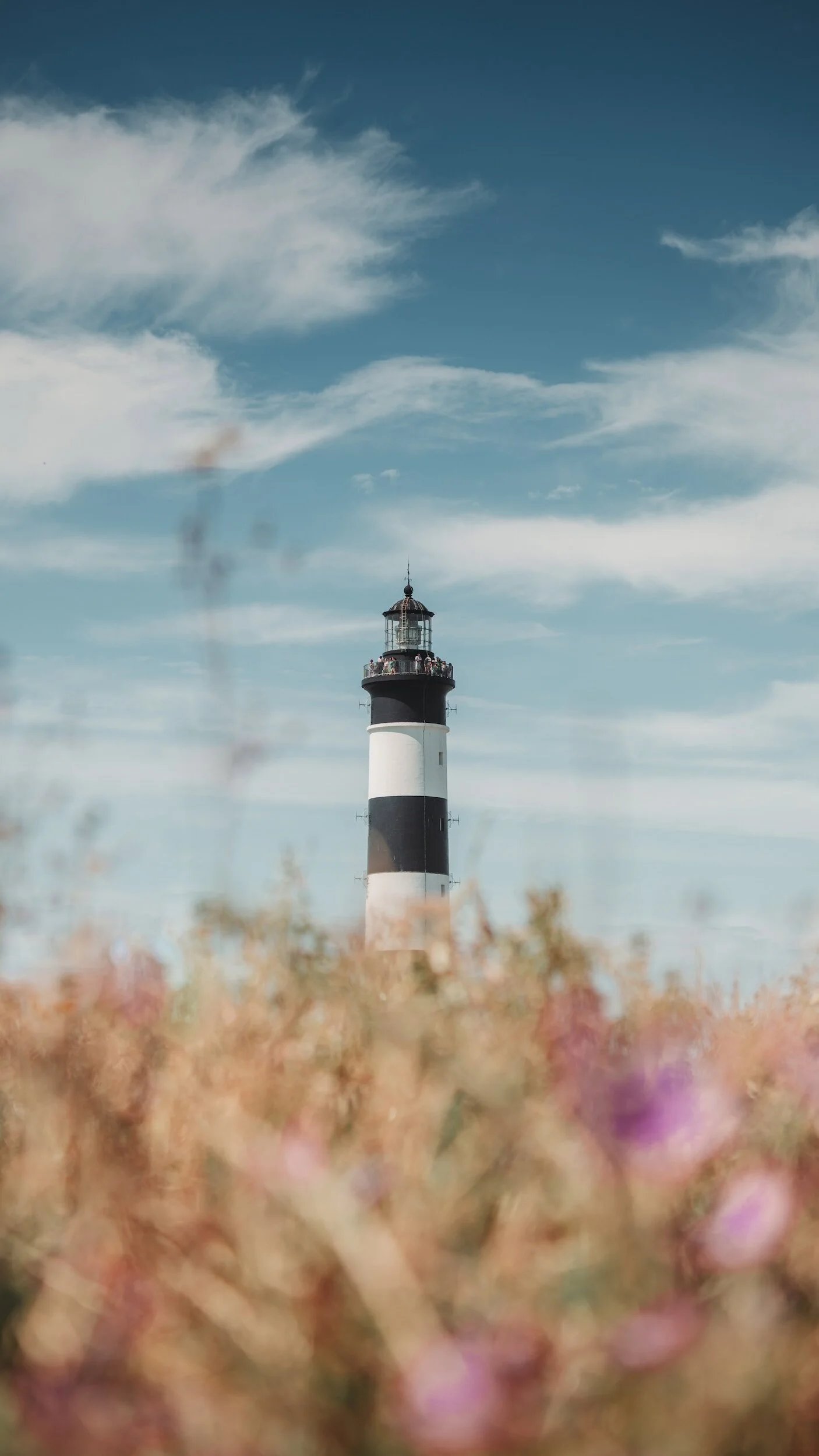 A lighthouse with black and white horizontal stripes standing against a partly cloudy sky with some clouds and blue sky.
