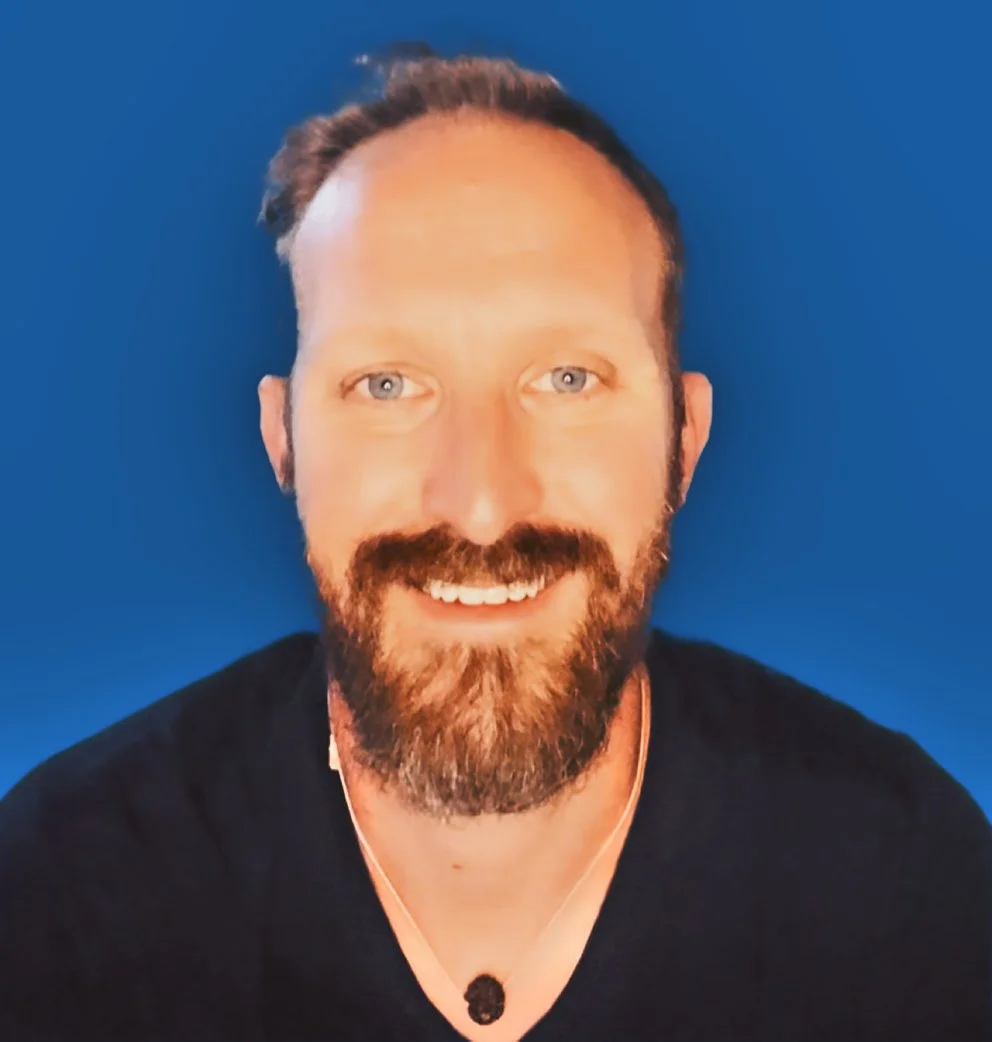 Portrait of a smiling man with a beard and mustache, wearing a black shirt and a necklace, against a blue background.