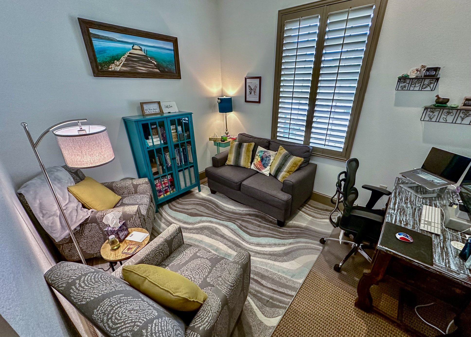 Cozy home office with two armchairs, a small sofa, a desk with a computer, bookshelf, wall art, and a window with blinds. The room features a patterned rug and a combination of modern and traditional decor elements.