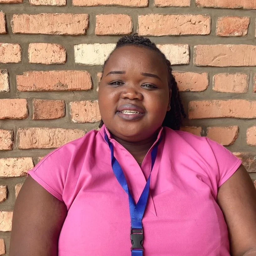 A smiling woman with dark skin tone wearing a short sleeve pink shirt and a blue lanyard around her neck