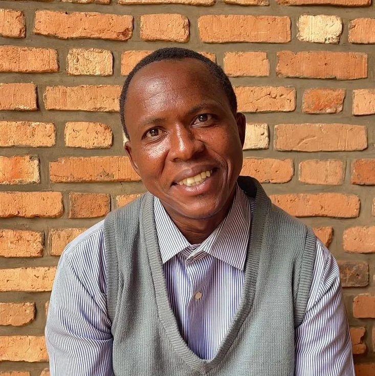 A smiling man with dark skin tone wearing a blue striped shirt and gray vest in front of a brick wall