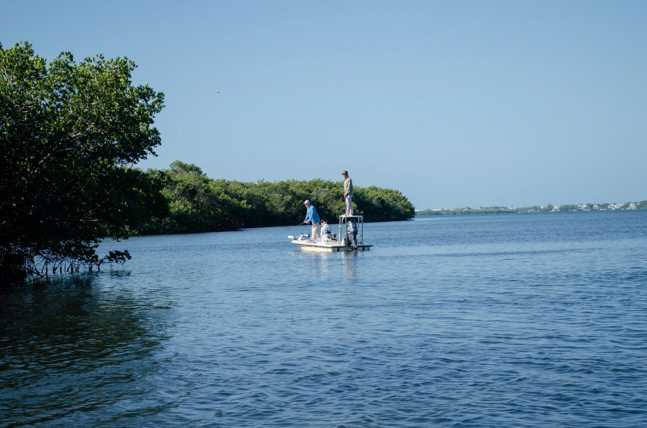 Two people fishing from a small boat on a calm body of water, with lush green trees along the shoreline and a clear blue sky.
