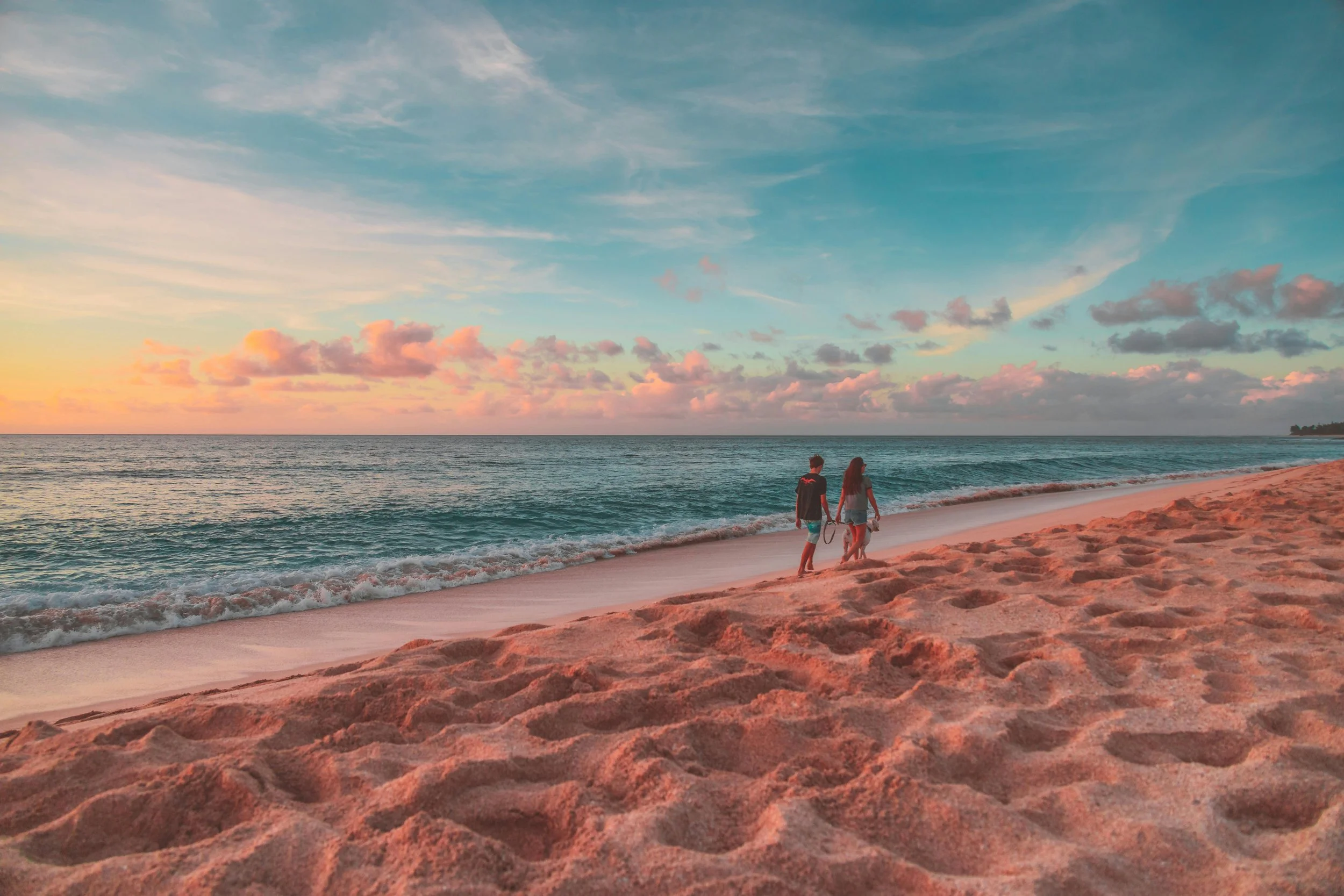 Two people walking along the beach near the water at sunset or sunrise, with a sky filled with soft pink and blue clouds.