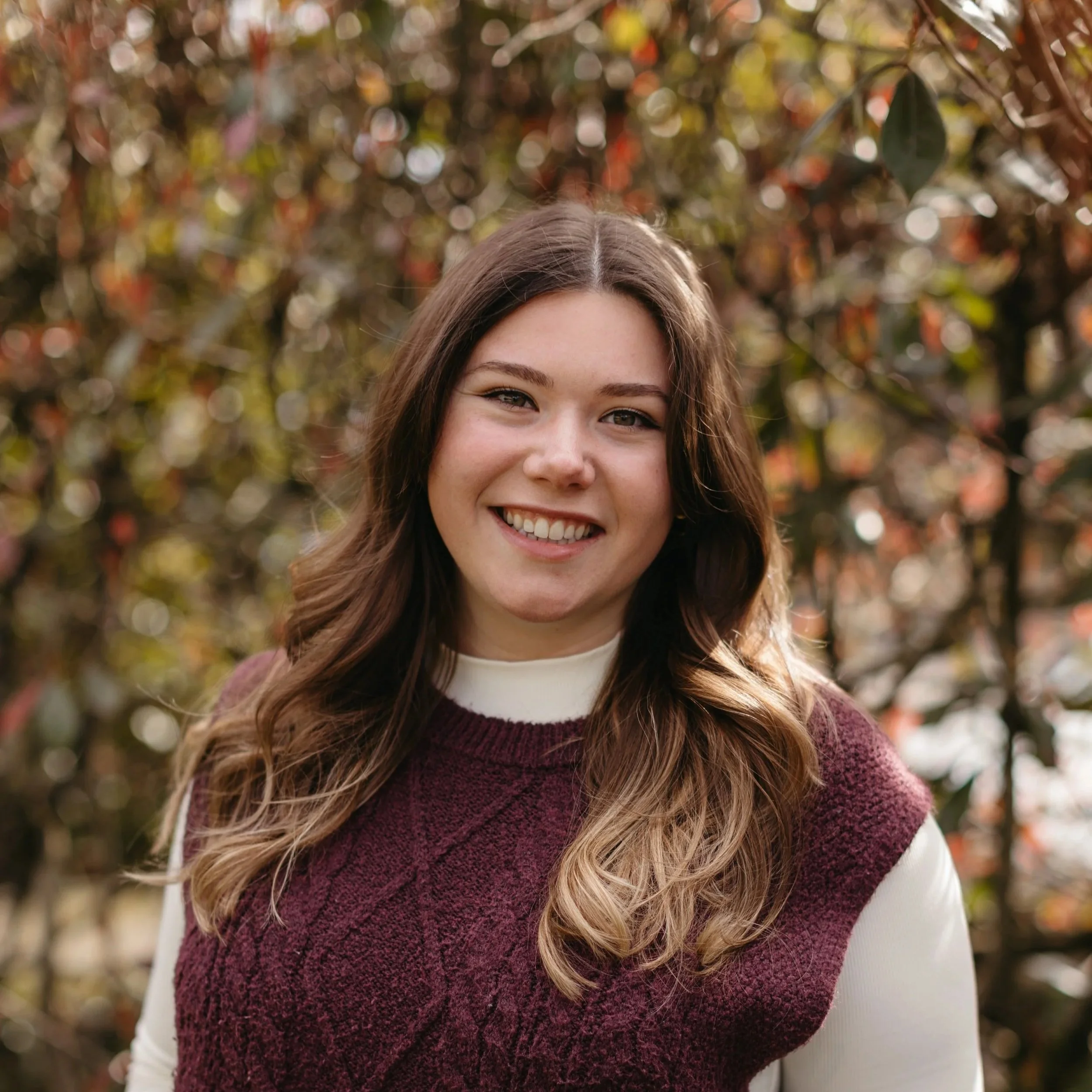 Close-up of a smiling young woman with wavy hair, wearing a white long sleeve shirt and a maroon vest, outdoors with blurred autumn leaves in background.