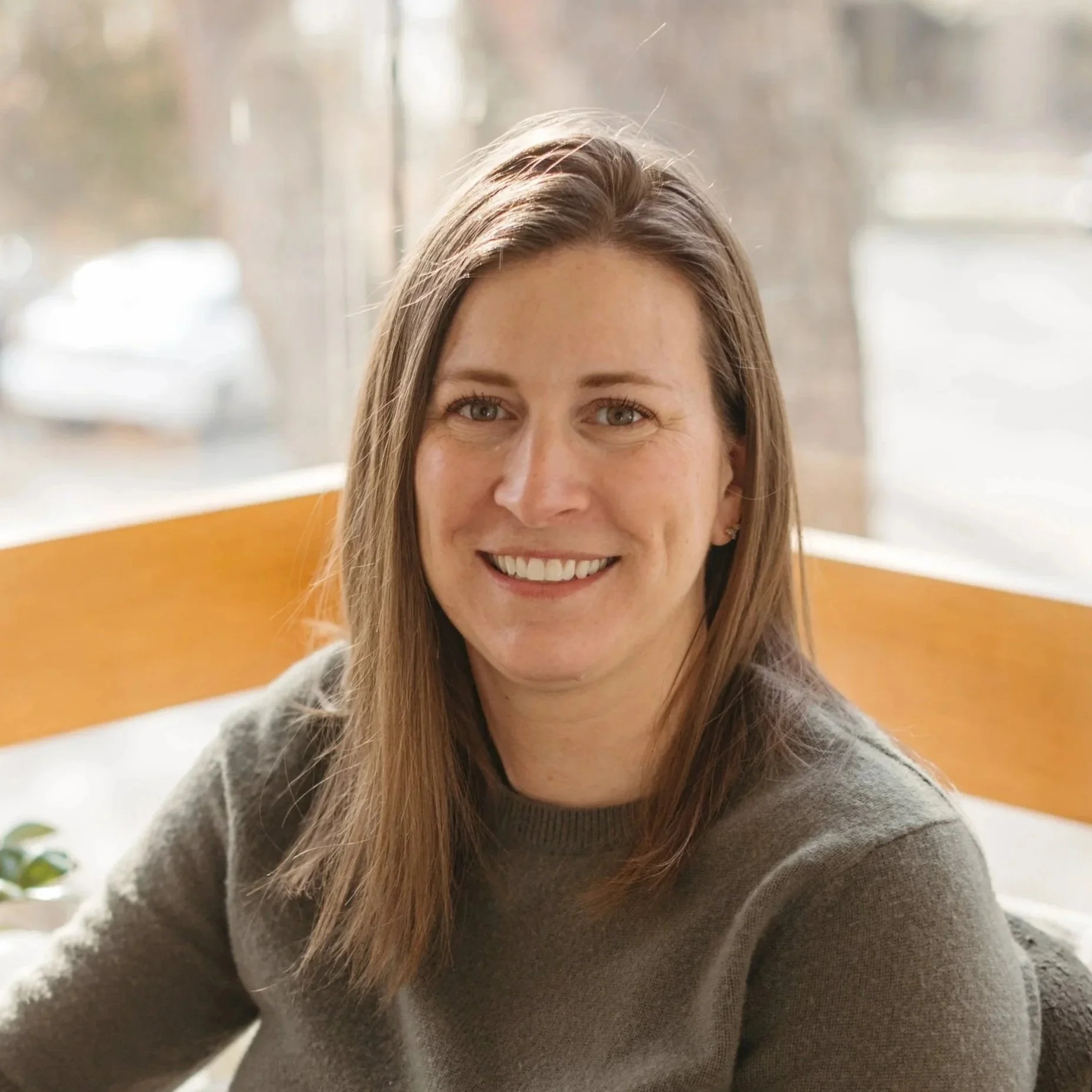 Close-up of a smiling woman with long brown hair, wearing a gray sweater, sitting indoors near a window with blurred background.