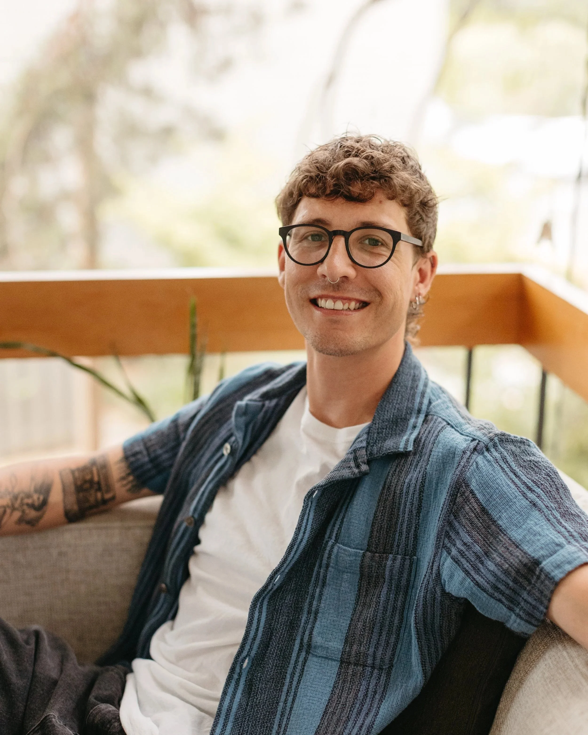 Close-up of a smiling person with short curly brown hair, wearing black rectangular glasses, a silver nose ring, and a gray striped sweater.