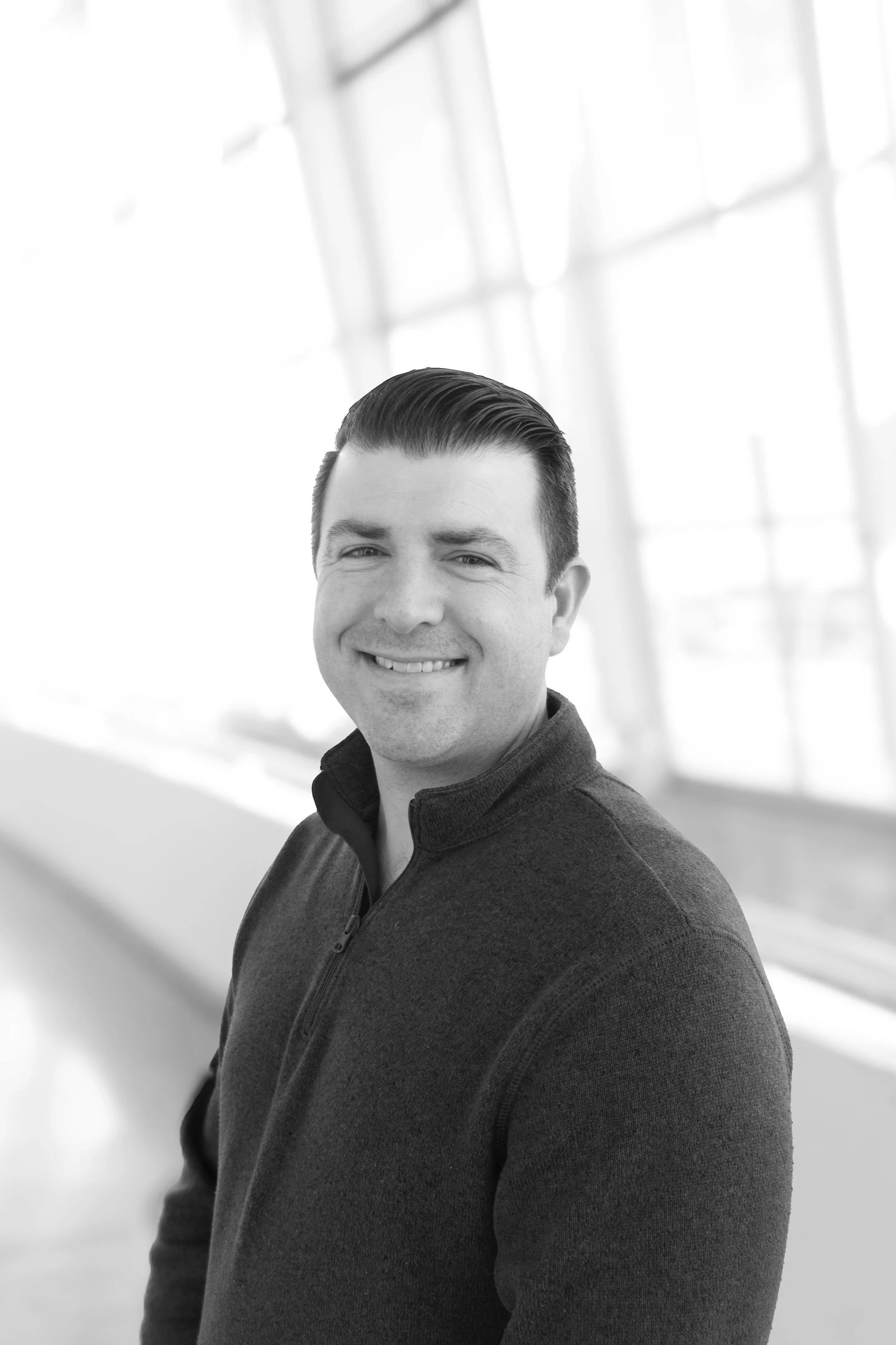 Black and white photo of a smiling man with dark hair slicked back, wearing a dark zip-up jacket, standing indoors with large windows in the background.