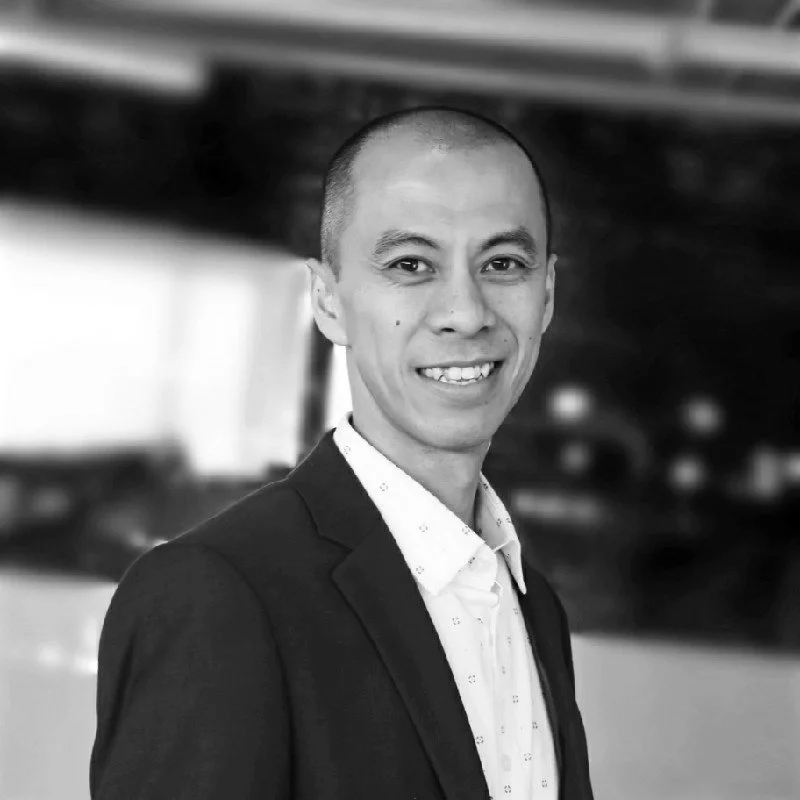 A smiling man with a shaved head wearing a suit jacket and collared shirt, standing indoors with a blurred background.