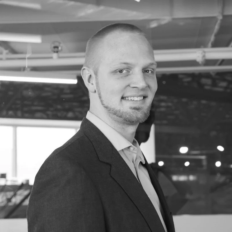 Black and white photo of a man with short hair, a goatee, wearing a suit jacket and collared shirt, smiling at the camera indoors.