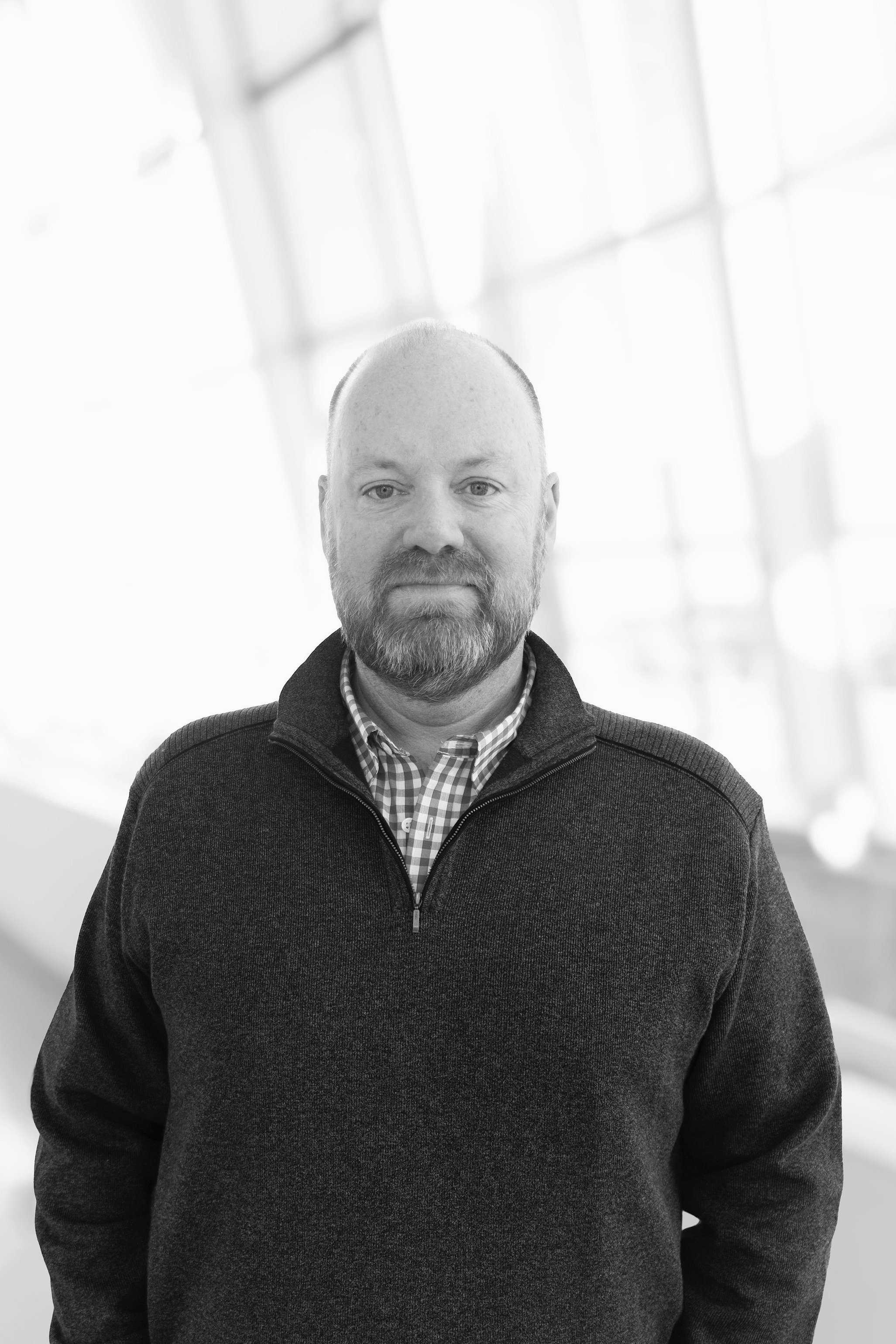 Black and white portrait of a man with a beard, wearing a checkered shirt and a zip-up sweater, standing inside a building with large windows in the background.