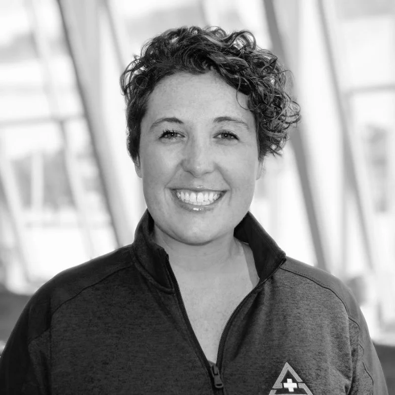 A woman with short, curly hair smiling, wearing a zip-up jacket with a sports logo, standing indoors near large windows.