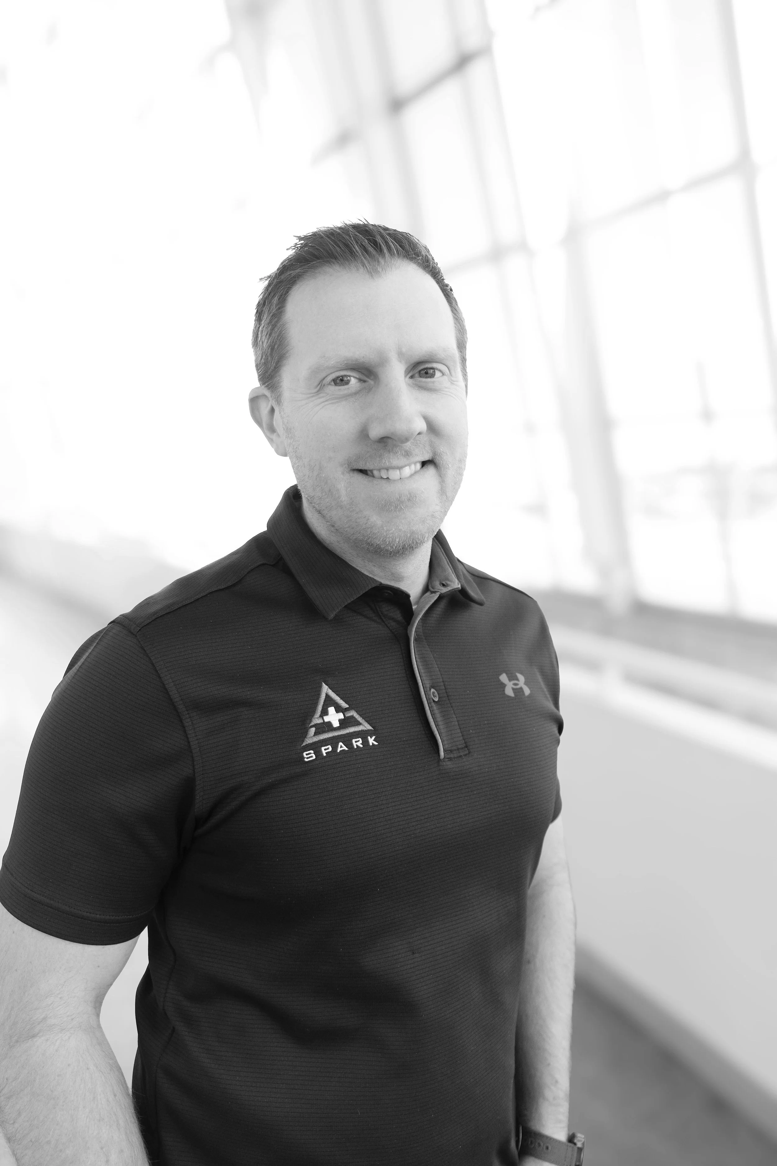 A smiling man in a black polo shirt standing indoors in front of large windows.