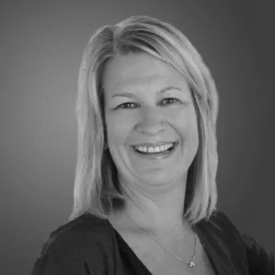 Black and white professional headshot of a smiling woman with shoulder-length hair, wearing a dark top and a necklace.