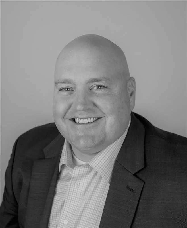 Black and white portrait of a bald man with a big smile, wearing a suit jacket and a checkered shirt, against a plain light background.