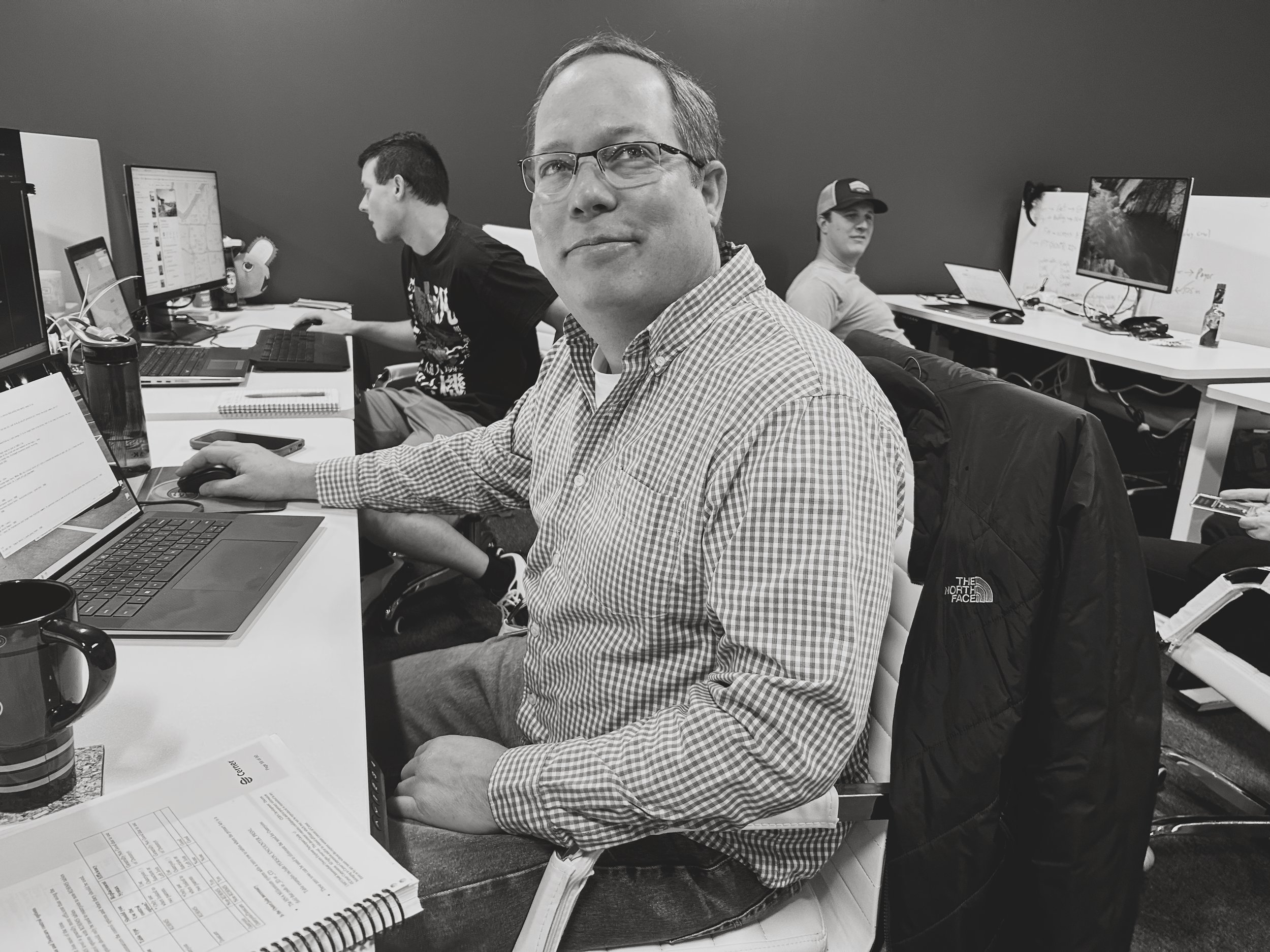 A man wearing glasses and a checkered shirt sitting at a desk with a laptop, in an office with two other men working at their computers in the background.