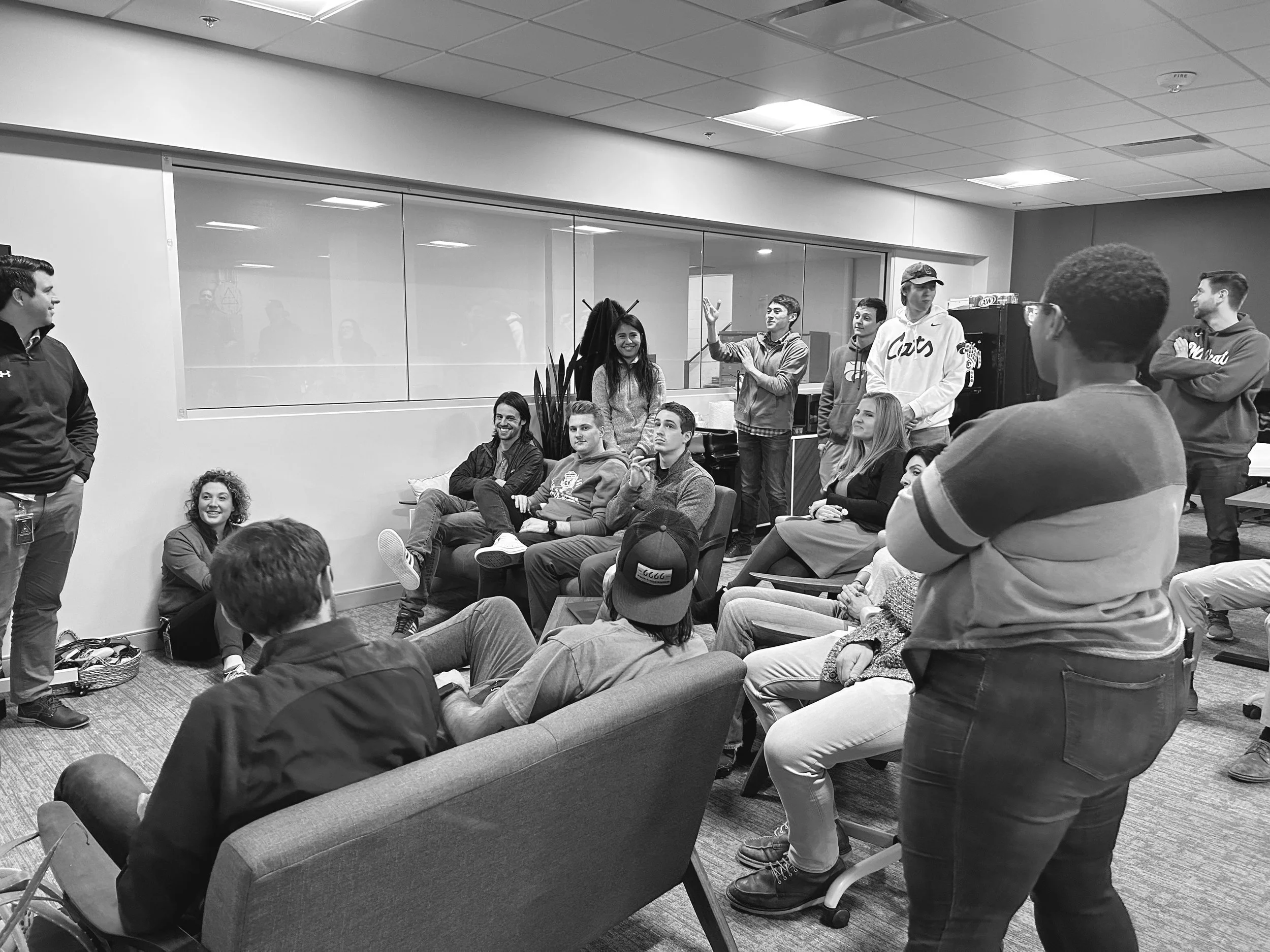 A group of young adults in a meeting room, some sitting and some standing, engaged in a discussion or presentation.