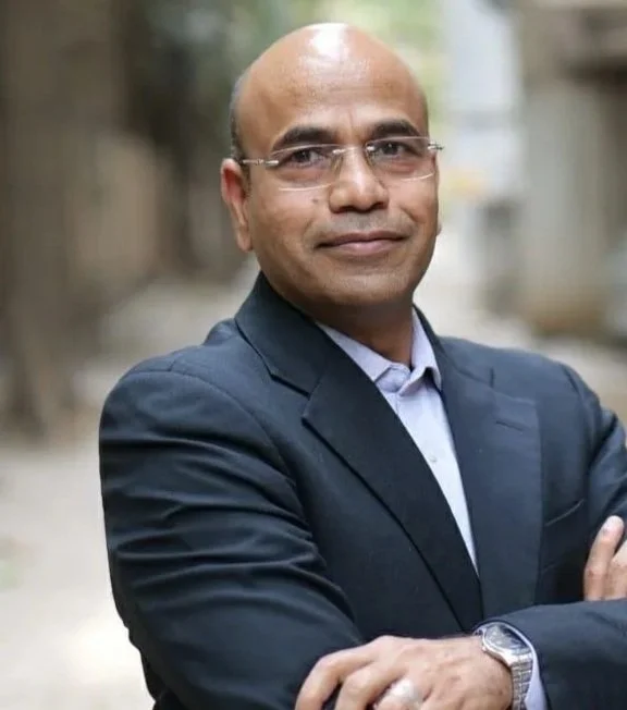 A man in a dark suit with glasses and a wristwatch, standing outdoors with arms crossed, in a blurred street background.