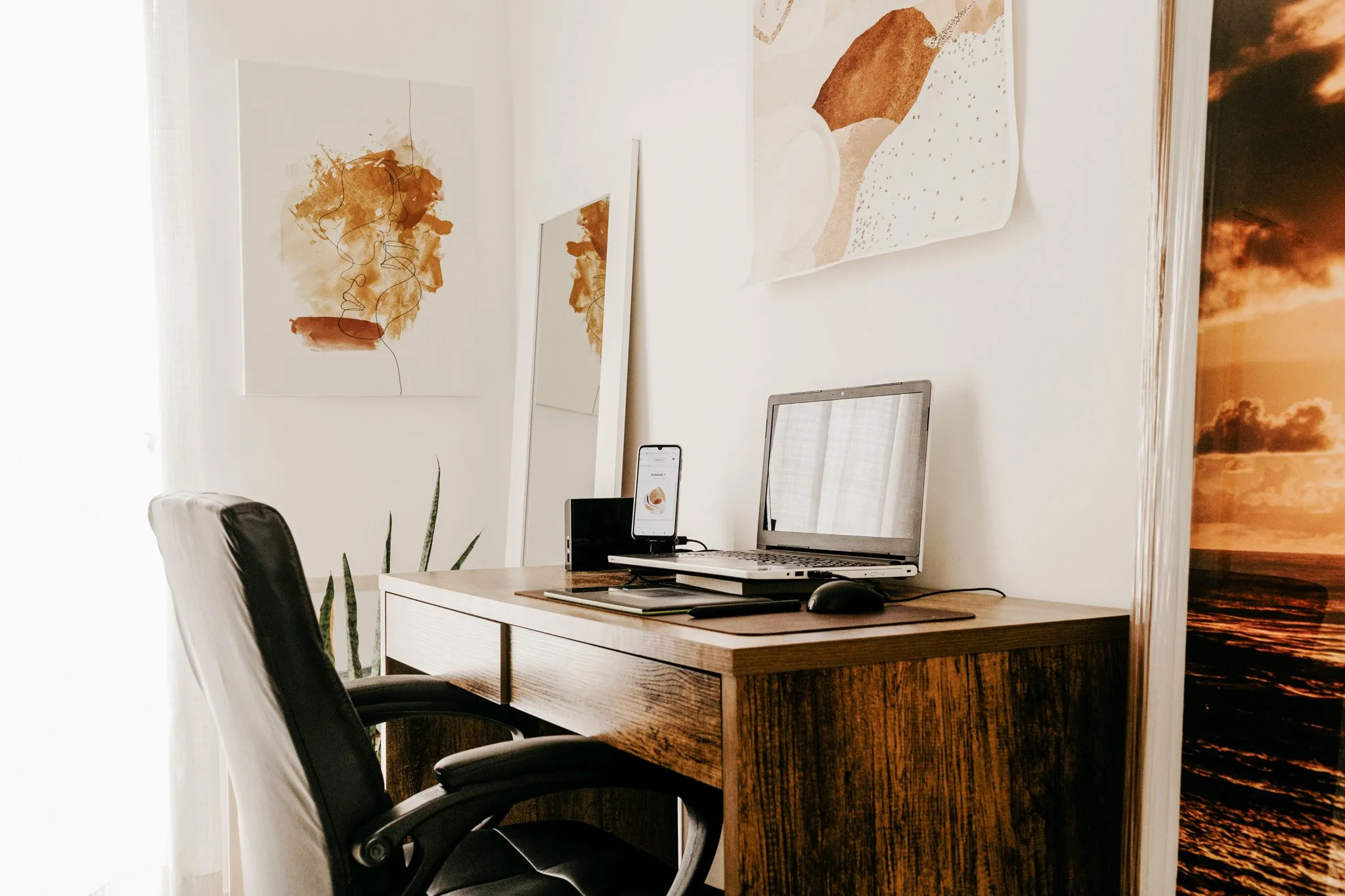 Home office setup with wooden desk, laptop, smartphone, mouse, and chair, decorated with abstract art and a sunset ocean poster on the wall.