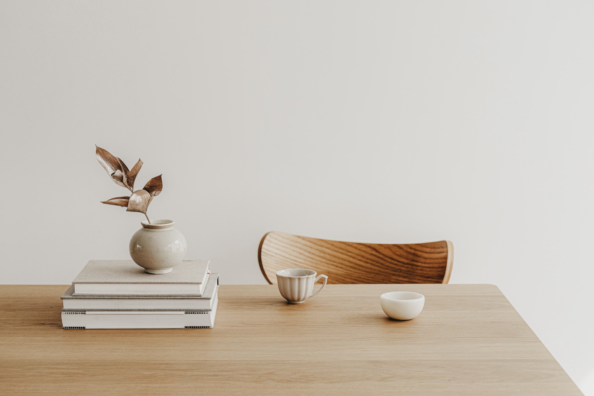 Minimalist dining table with a stack of books, a beige vase with dried leaves, a striped teacup, and a small white bowl, against a plain white wall.