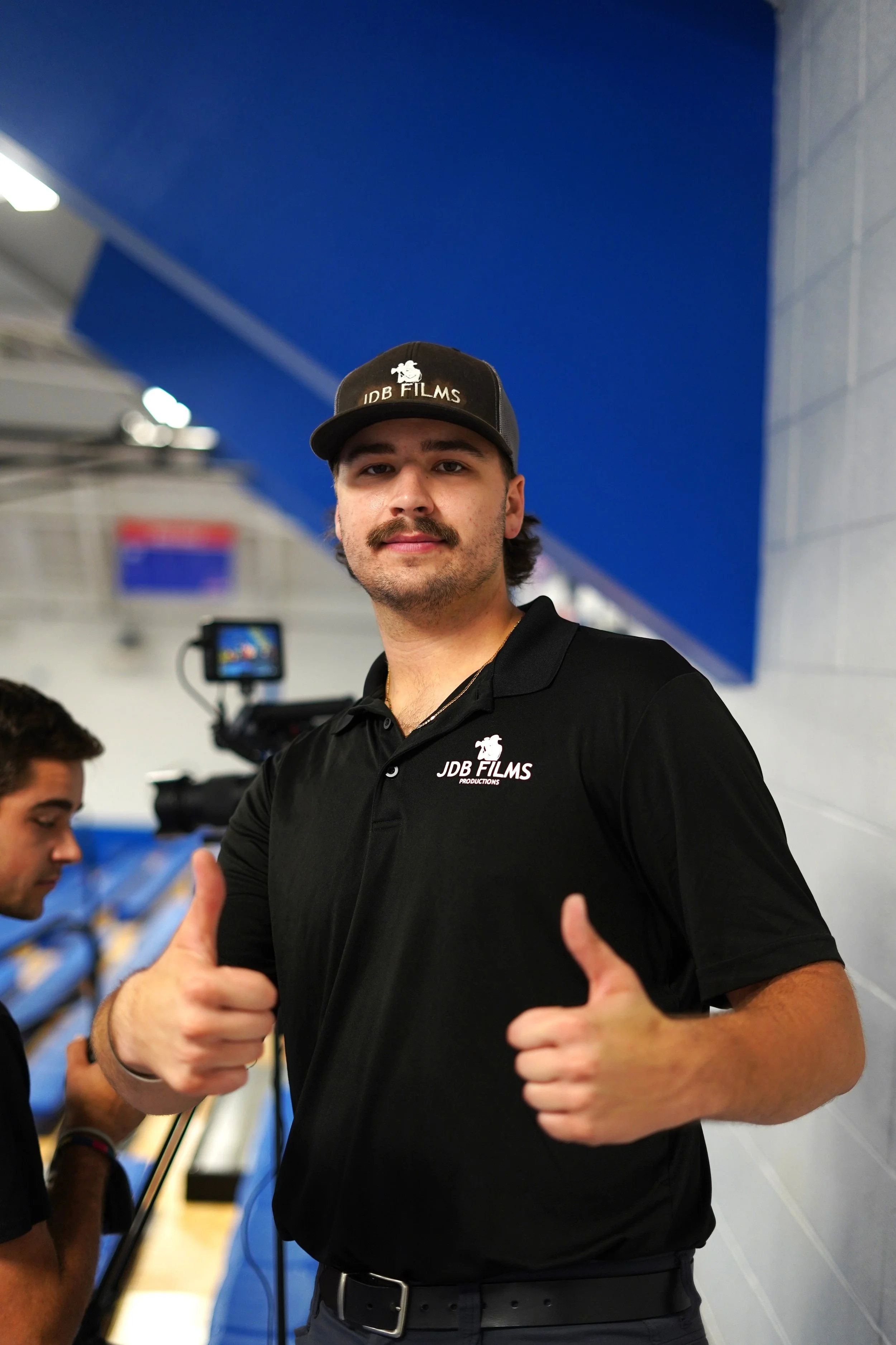 A man in a black polo shirt and cap with the logo 'JDB FILMS' gives a thumbs up inside a gymnasium with bleachers and a camera in the background.