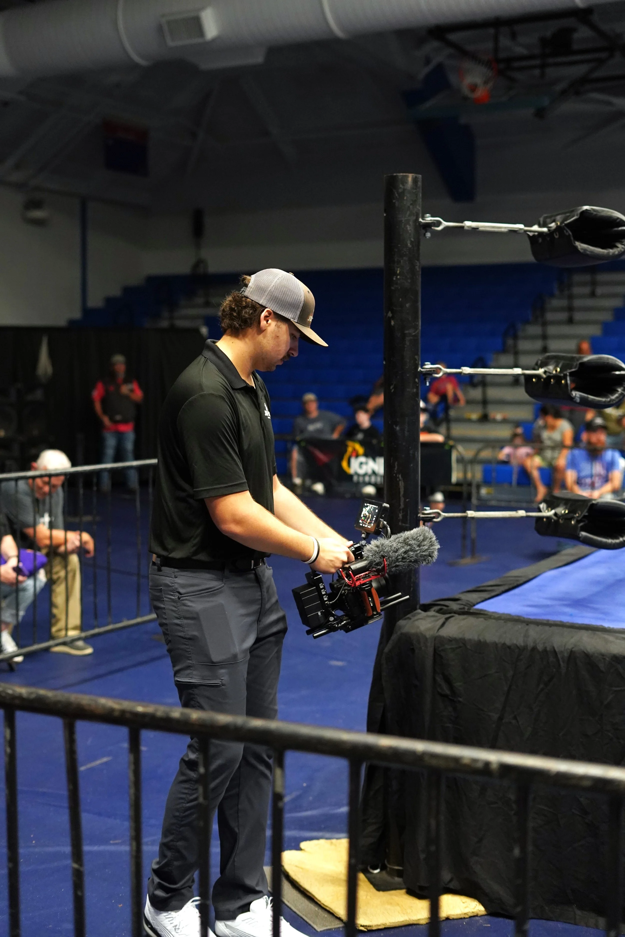 A man operating a professional video camera with a microphone attached, standing next to a wrestling ring at an indoor sports arena.