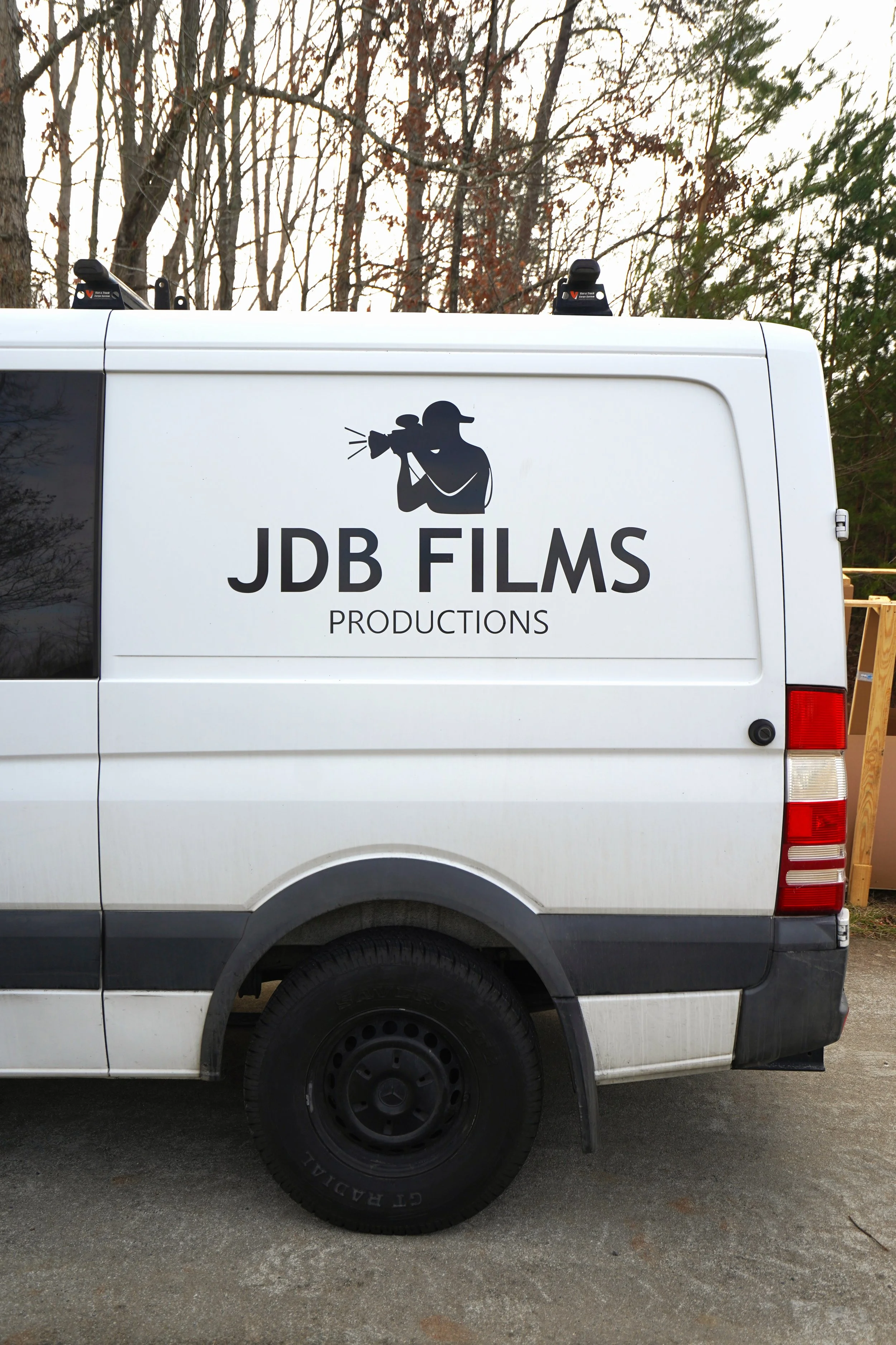 A white van with the logo of JDB Films Productions, featuring a silhouette of a photographer with a camera and a hat, parked outdoors with trees in the background.