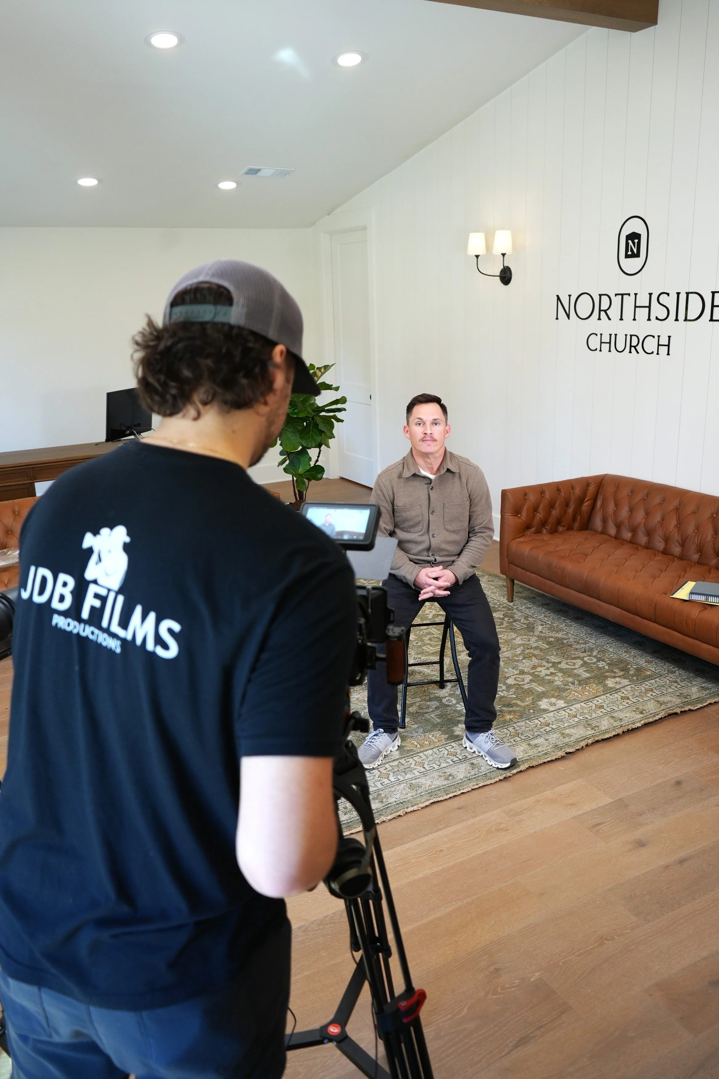 A man is being filmed during an interview in the lobby of Northside Church. The interviewer operates a camera, and the man is sitting on a stool, facing the camera, in front of a white wall with the church's logo.