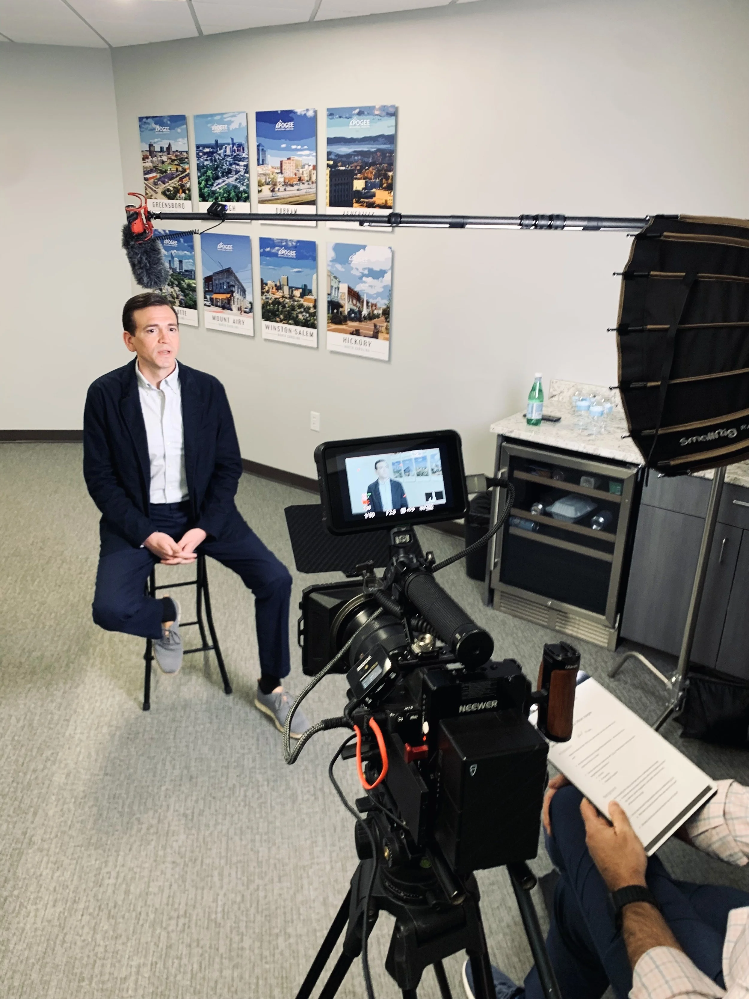 A man in a dark jacket and white shirt is seated on a stool in front of a camera during an interview or recording session in an office with framed cityscape posters on the wall. A person holding a booklet is partially visible on the right.