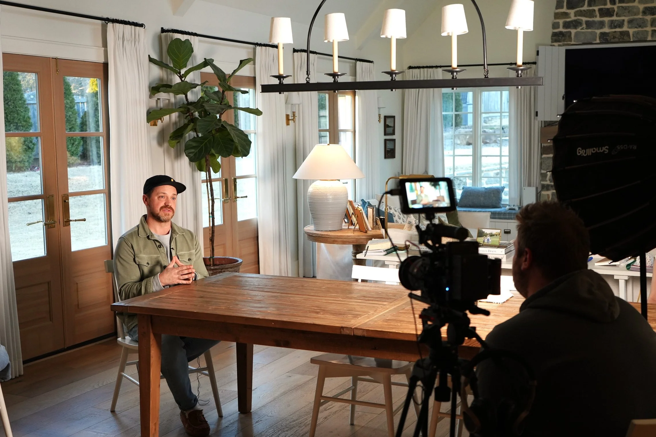 A man being filmed during an interview in a cozy, well-lit living room with large windows, white curtains, a wooden table, a tall potted plant, a table lamp, and a camera operator.