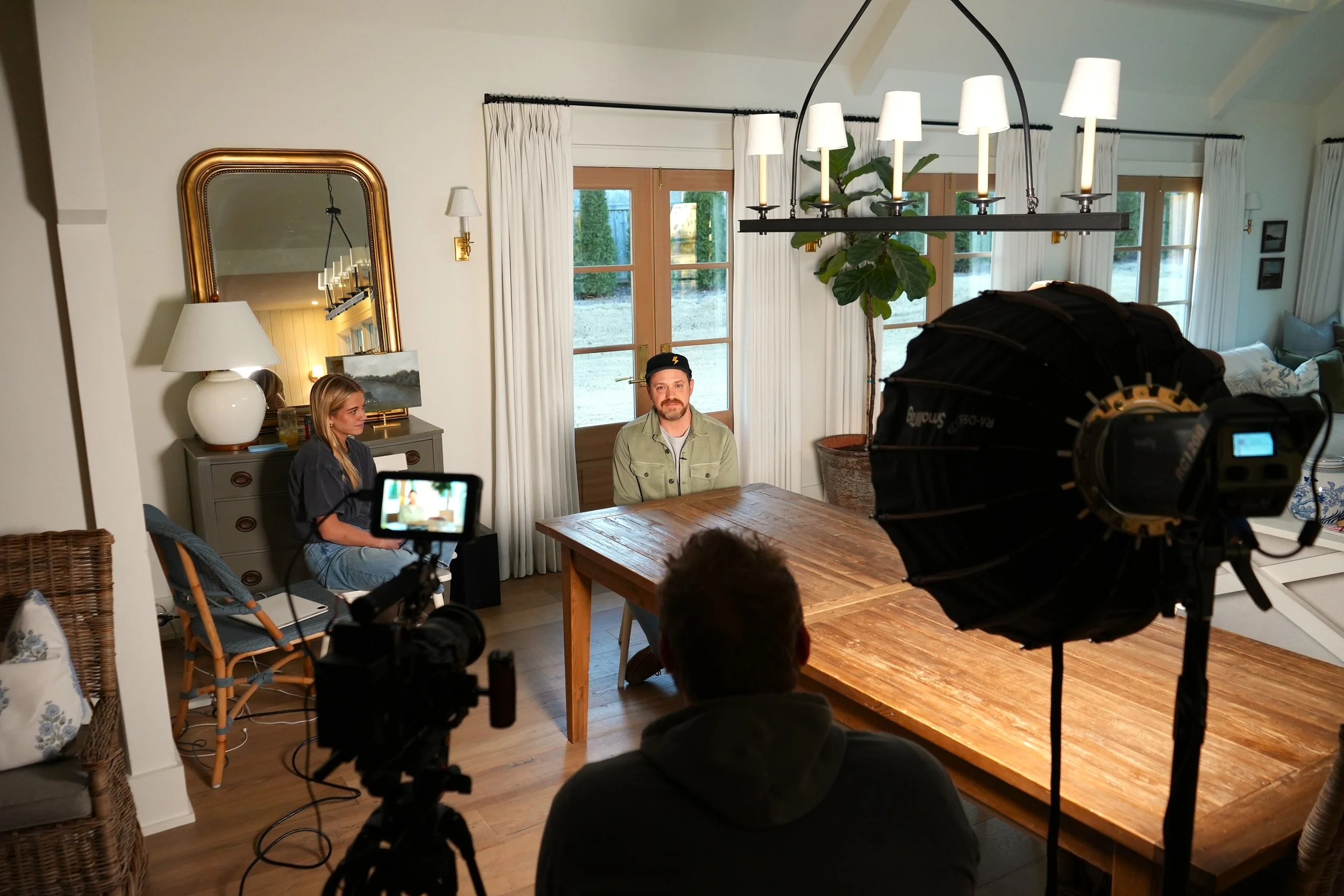 People filming an interview or scene in a cozy, well-lit room with wooden floors, white curtains, a large wooden table, and a leafy plant. Two camera operators and a director are visible, with one man seated at the table and a woman sitting nearby.