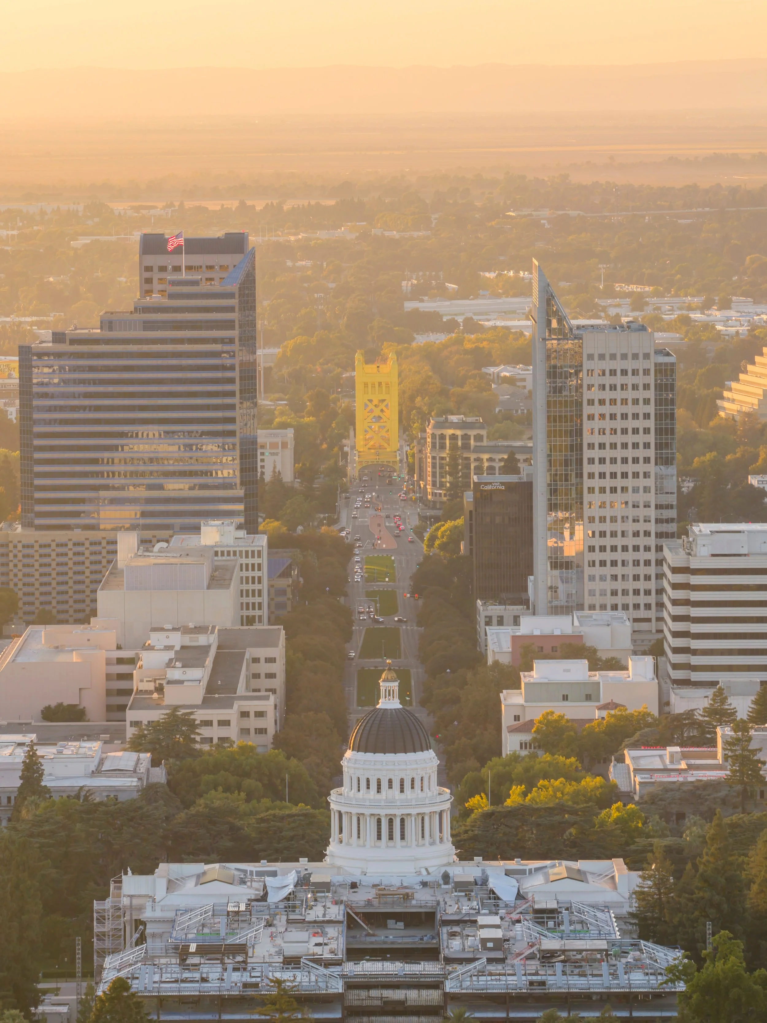 A city skyline at sunset with a prominent white capitol building with a dome in the foreground, and tall modern glass skyscrapers in the background, with trees and a hazy sky.