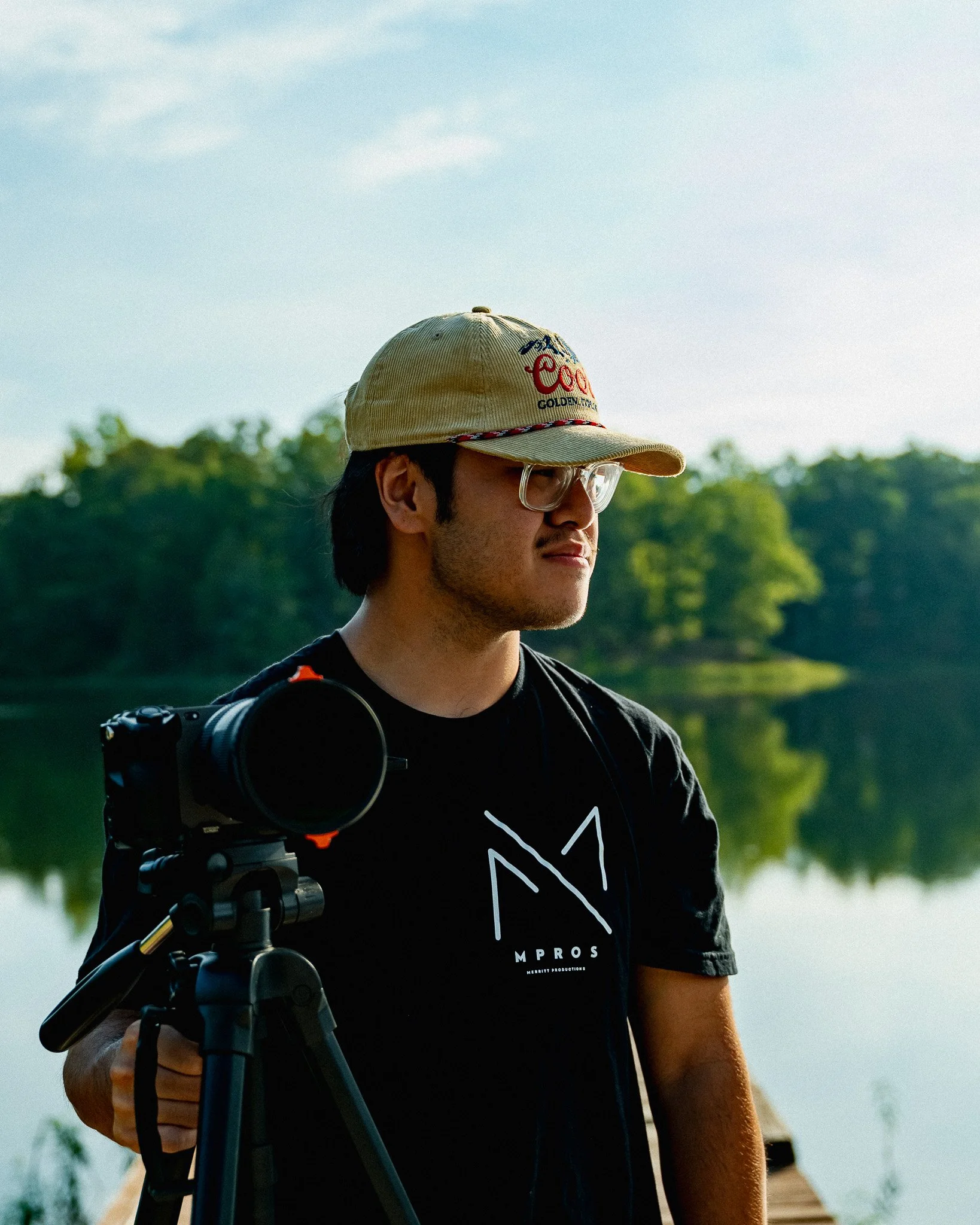 A man wearing glasses and a beige baseball cap with a logo, standing outdoors near a body of water with trees in the background, holding a camera on a tripod.