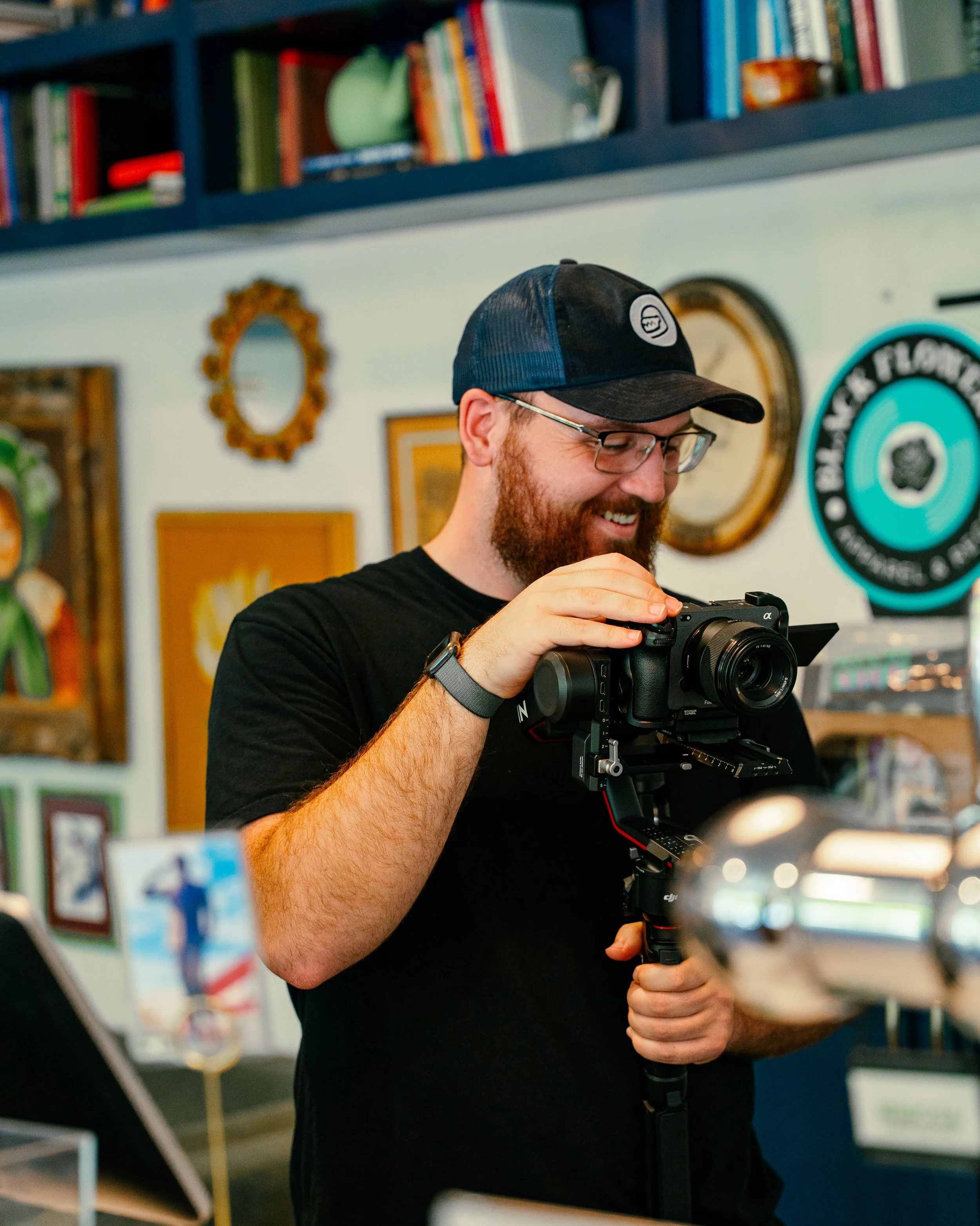 A man with glasses and a beard, wearing a black baseball cap and black t-shirt, is smiling while adjusting a professional camera on a stabilizer in a room decorated with bookshelves and wall art.