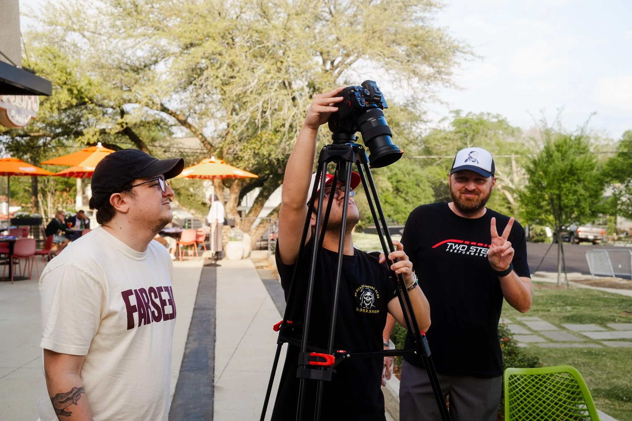 Three people participating in a video or photo shoot outdoors. One person is adjusting a camera on a tripod, and another is making a peace sign. The third person is observing. They are in a park-like setting with trees, umbrellas, and outdoor seating.