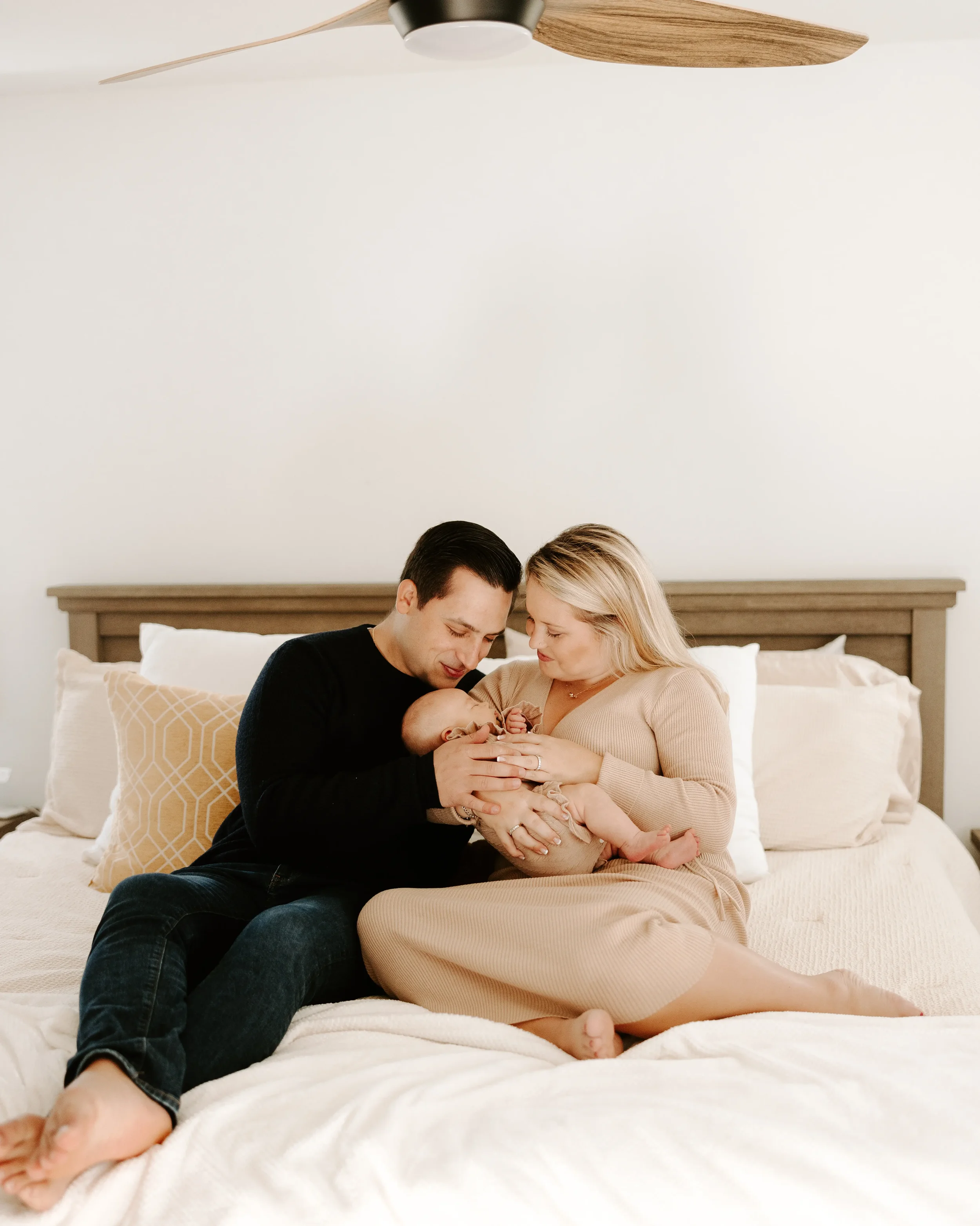 A couple sitting on a bed with a newborn baby, holding and looking at the baby lovingly in a well-lit bedroom.