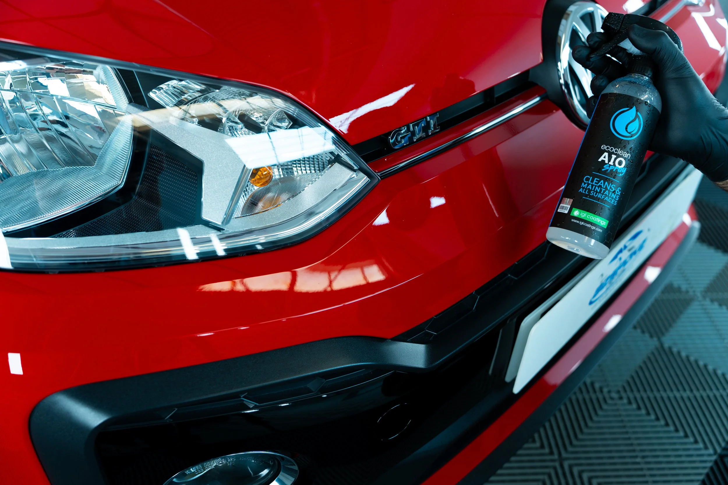 Close-up of a red car's front end, showing the headlight and a gloved hand holding a spray bottle labeled 'eco clean A10 Spray' near the hood.
