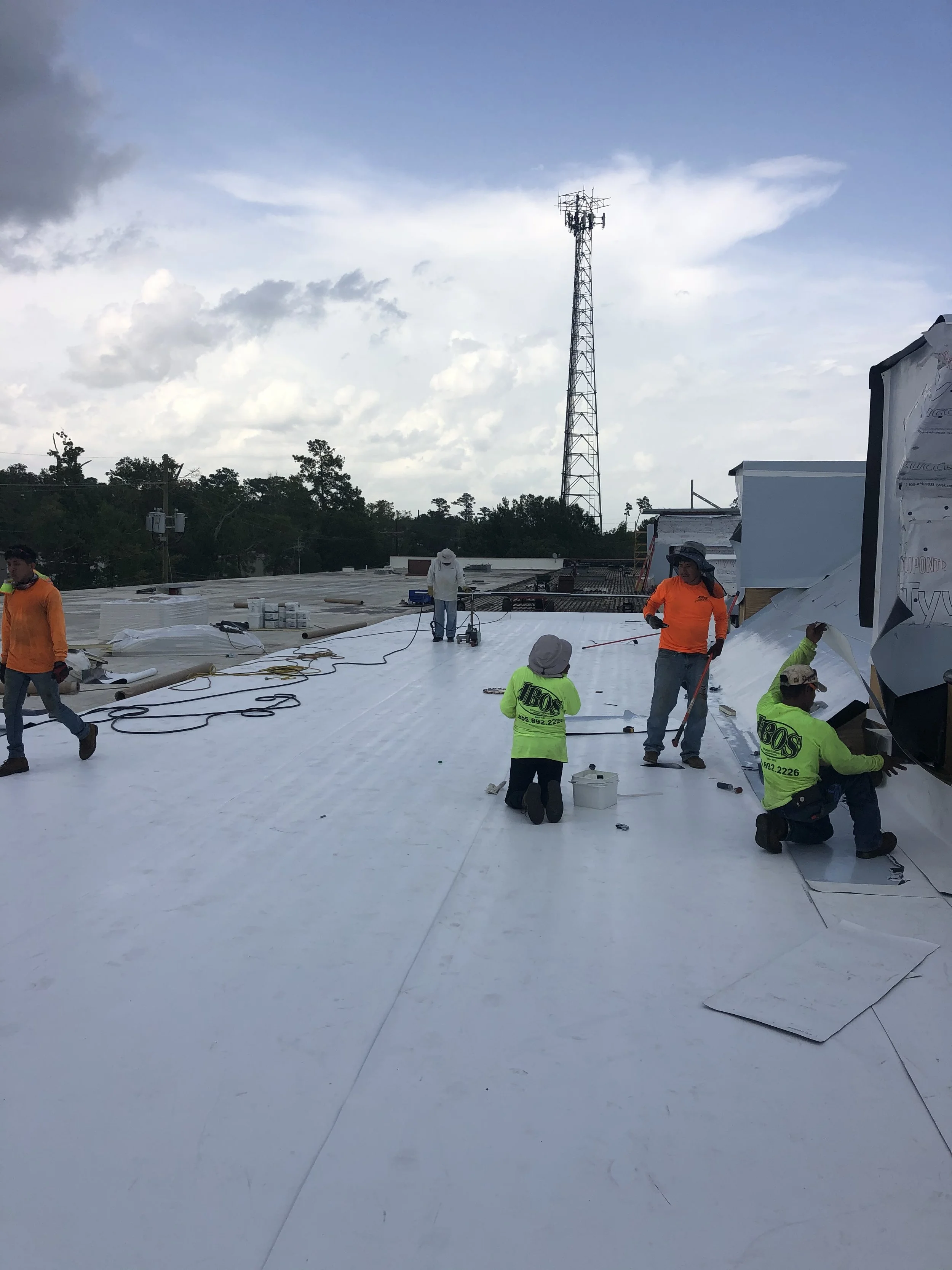 Construction workers installing a white roofing membrane on a flat rooftop during daytime, with a cloudy sky and a tall communication tower in the background.