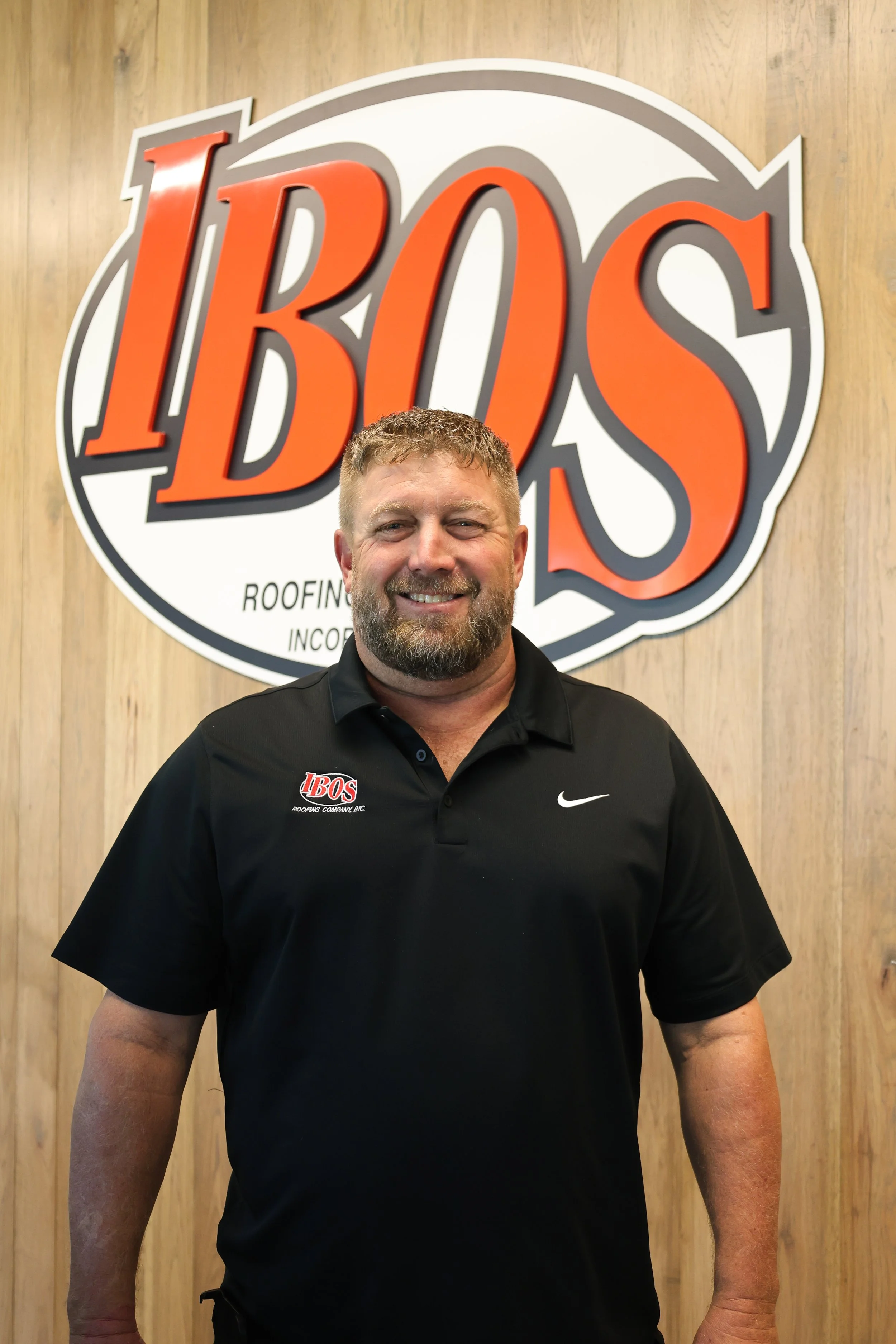 A smiling man with a beard wearing a black Nike polo shirt standing in front of a building with a sign that reads 'COMPANY' and has a logo with red and black initials.