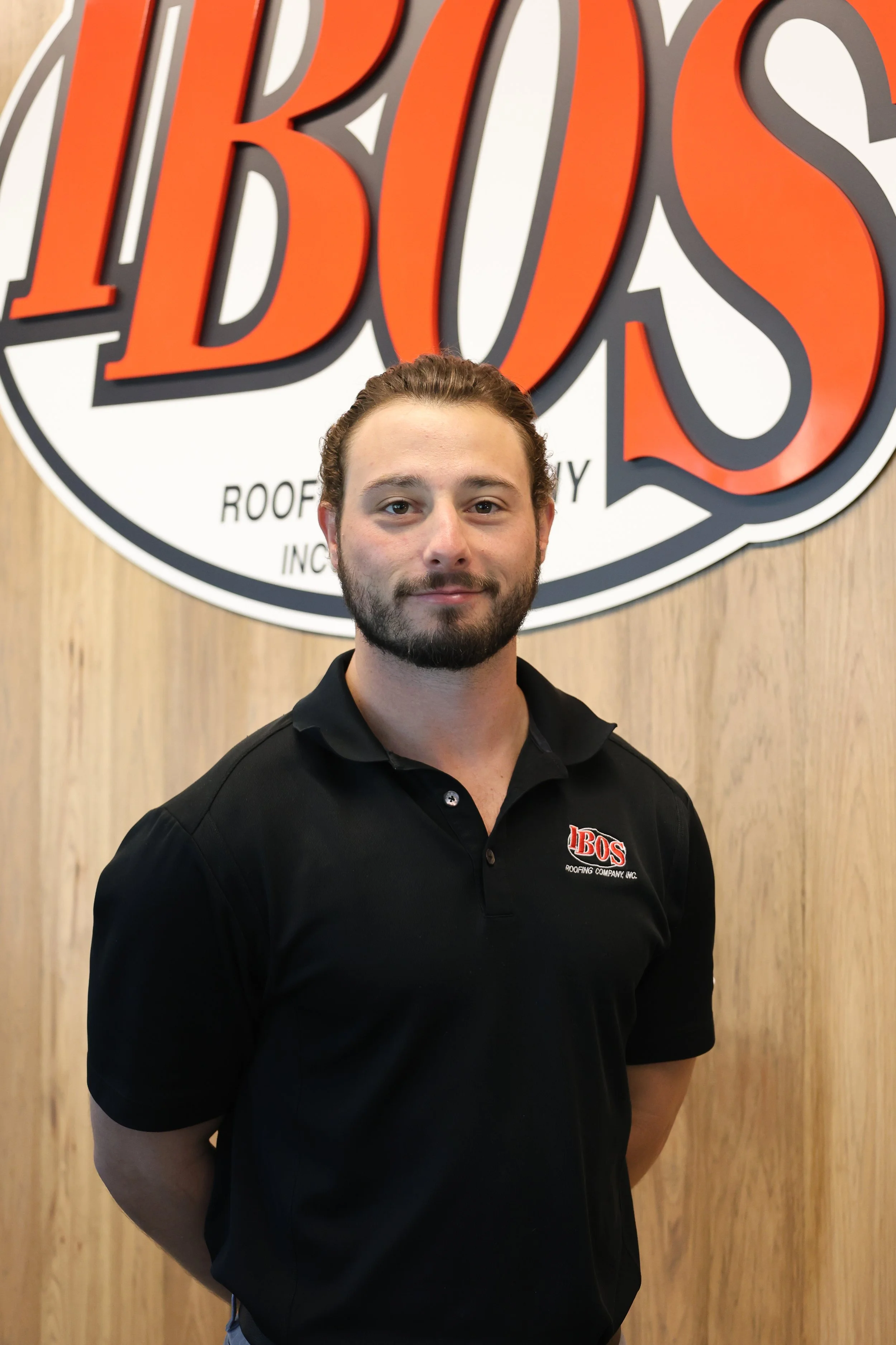 A smiling man in a black polo shirt with the logo 'IbOS Roofing Company Inc.' standing outdoors next to a wooden wall and a white sign with partial logo and black text that reads 'COMPANY'.