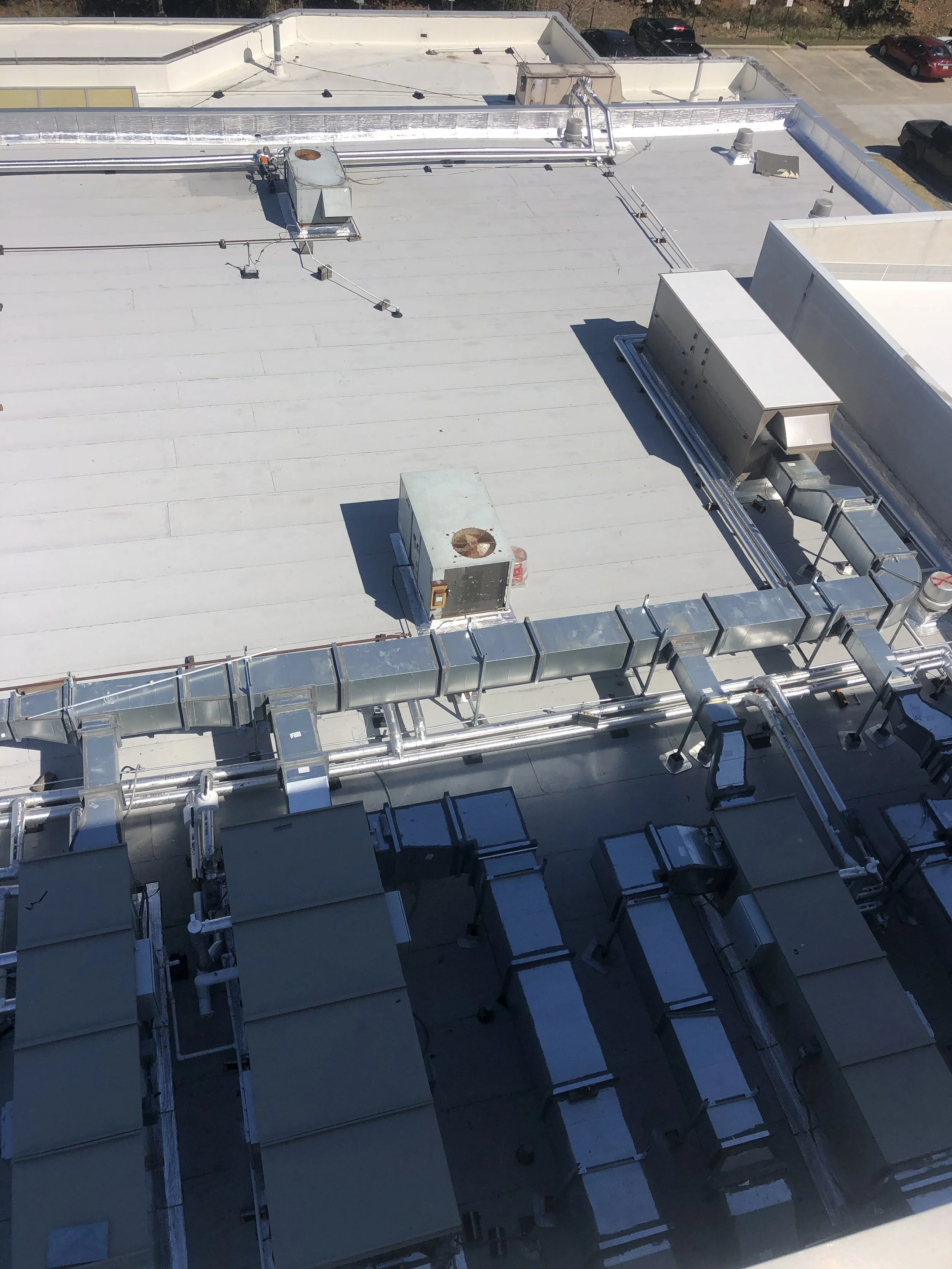 View of a building roof with HVAC units, ductwork, and electrical conduits, with a parking lot and some cars in the background.
