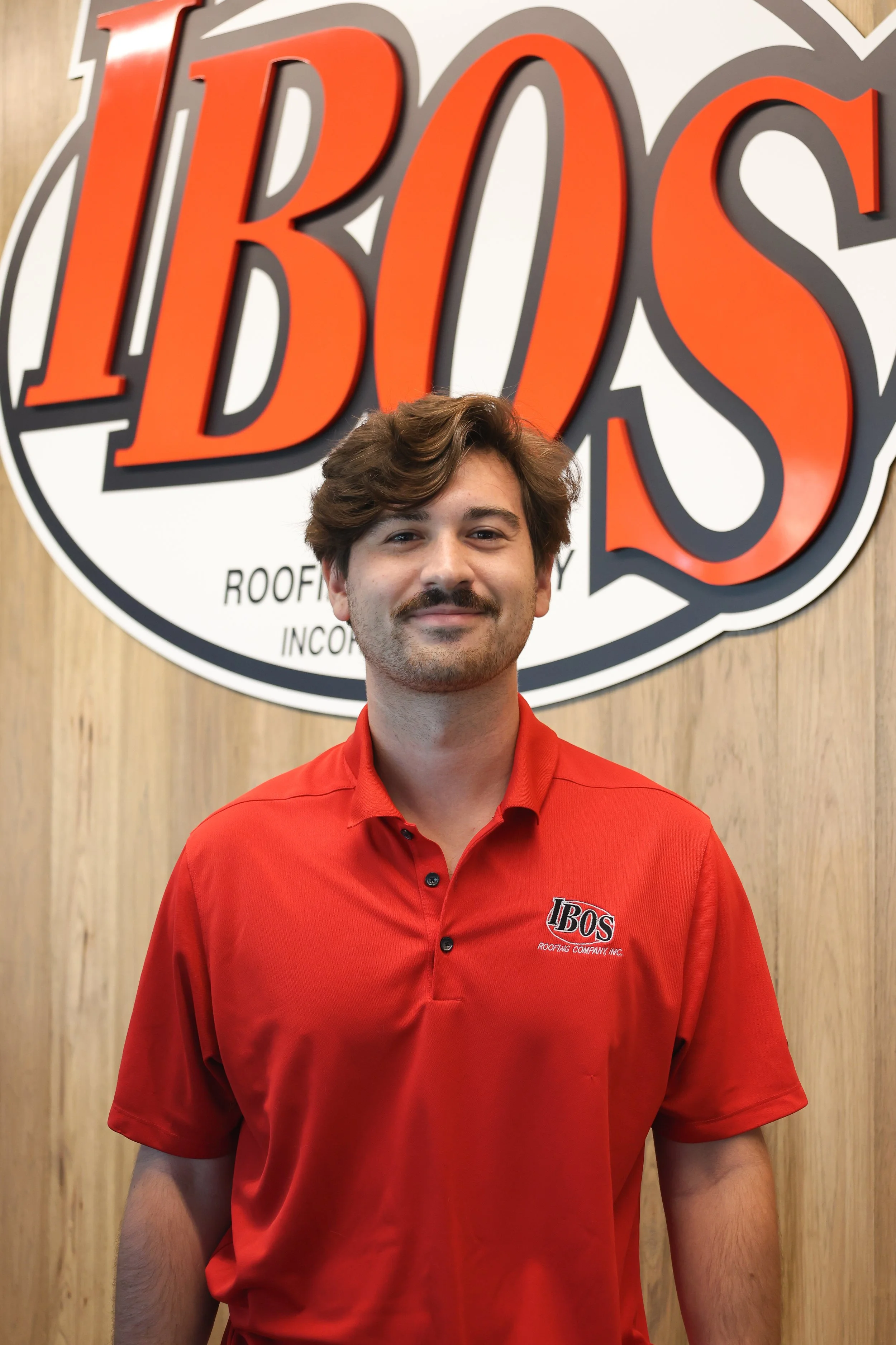 Young man with brown curly hair and beard, wearing a black polo shirt with a company logo, standing outside near a building with a sign that reads "IBOS" and part of another sign visible.