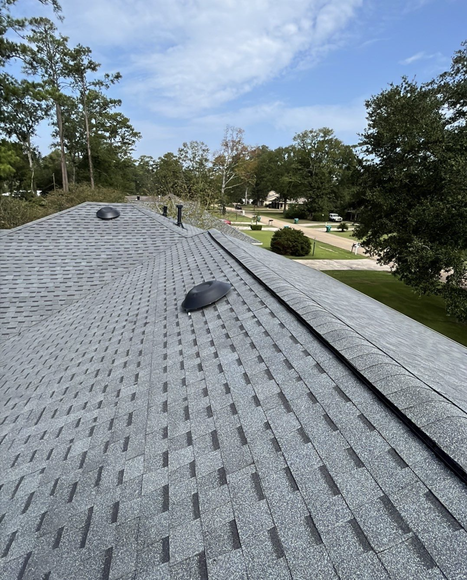 View of a gray shingle roof with two black roof vents, in a suburban neighborhood with trees and houses visible in the background under a partly cloudy sky.