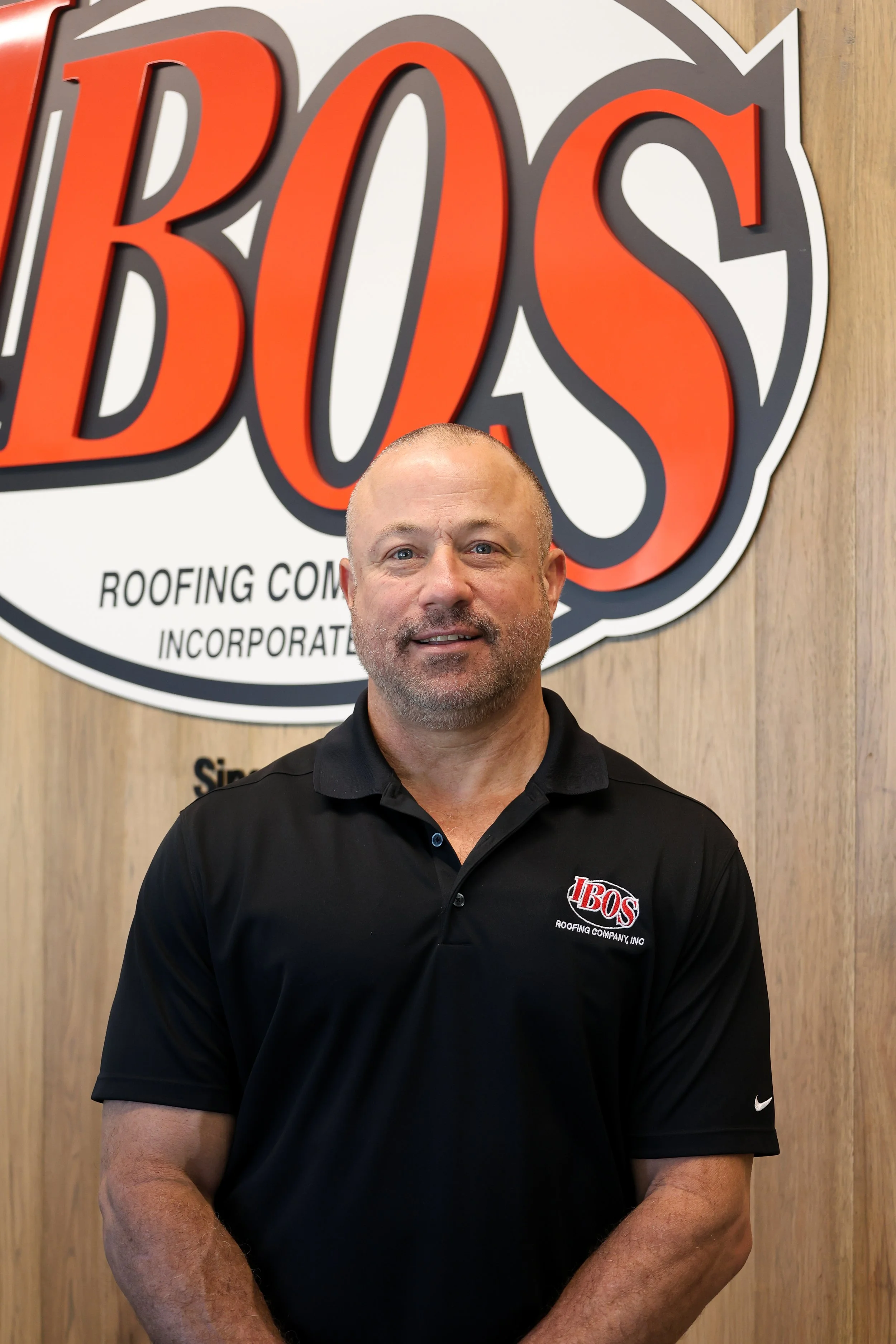 A man wearing a black polo shirt with a logo on it, standing in front of a sign with the word 'BOS' in large red letters. The sign also has smaller text underneath that is partially visible.