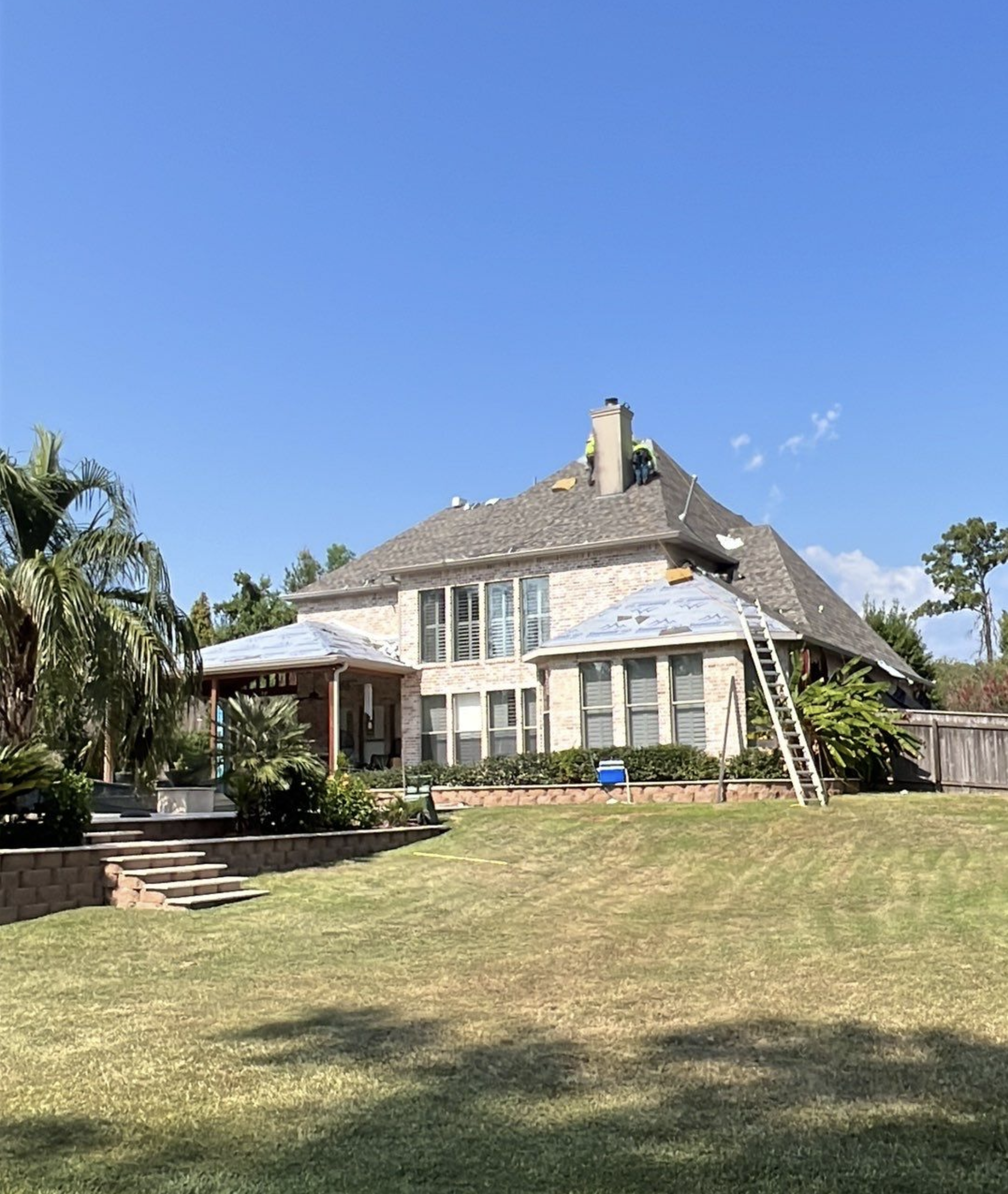 A large two-story brick house with a shingled roof under construction or repair, with ladders leaning against it, and a well-maintained lawn and trees in the yard.