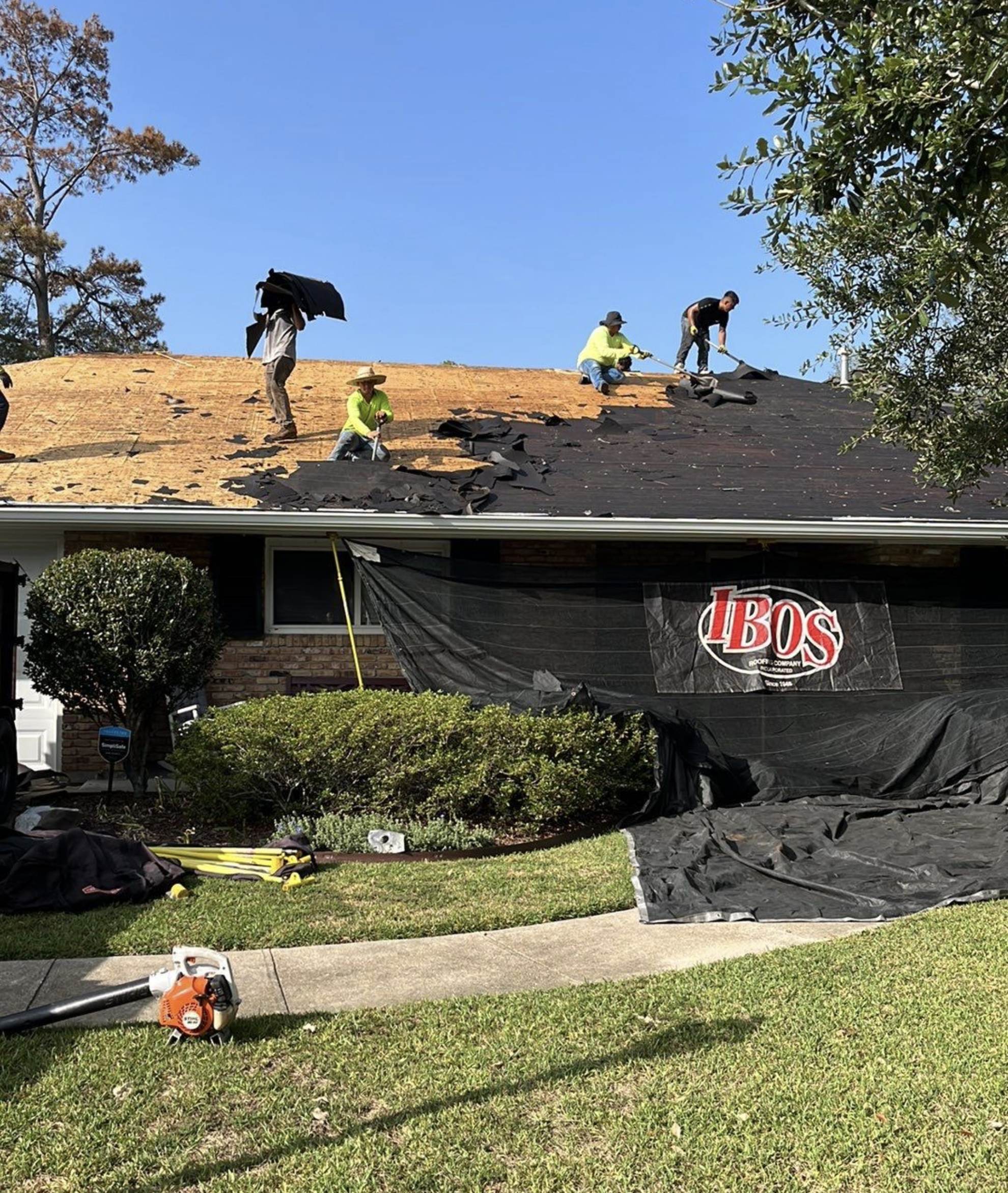 Construction workers repairing or replacing the roof of a house. Some workers are on the roof, removing old material, while others are working with tools. The house has a brick exterior, with a front yard and bushes, and a ladder is leaning against the house.