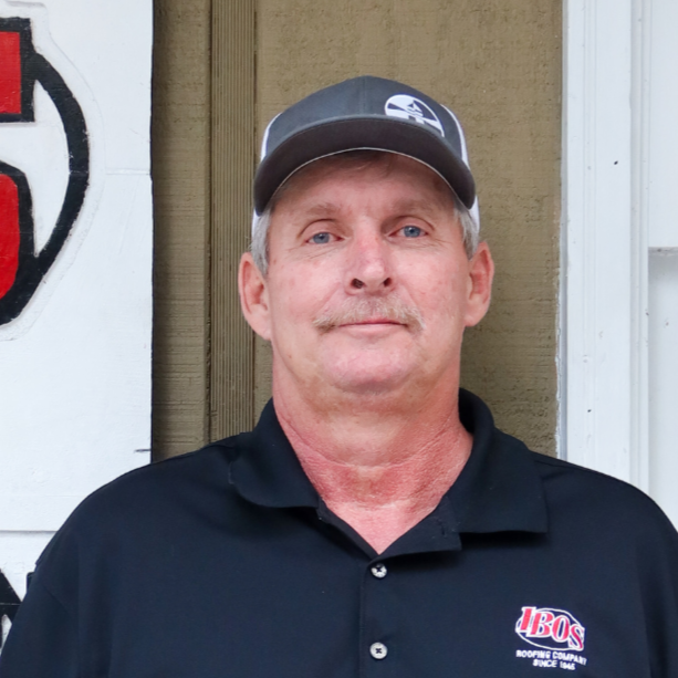 A man wearing a black polo shirt and a baseball cap standing outside next to a sign for a roofing company named IBOS.