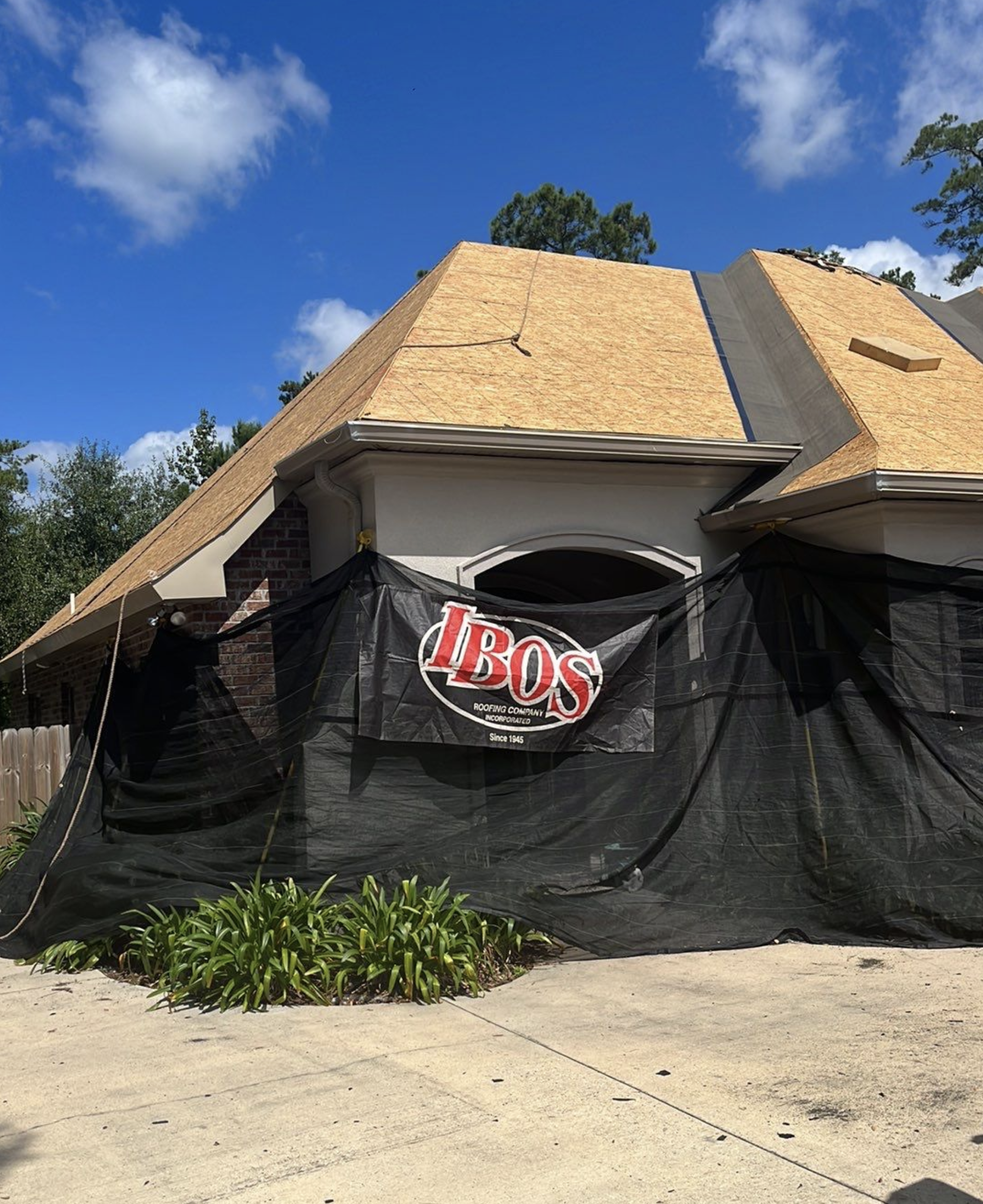 House under construction with a new roof being installed, covered with black construction netting, and a banner from Ibos Roofing Company.